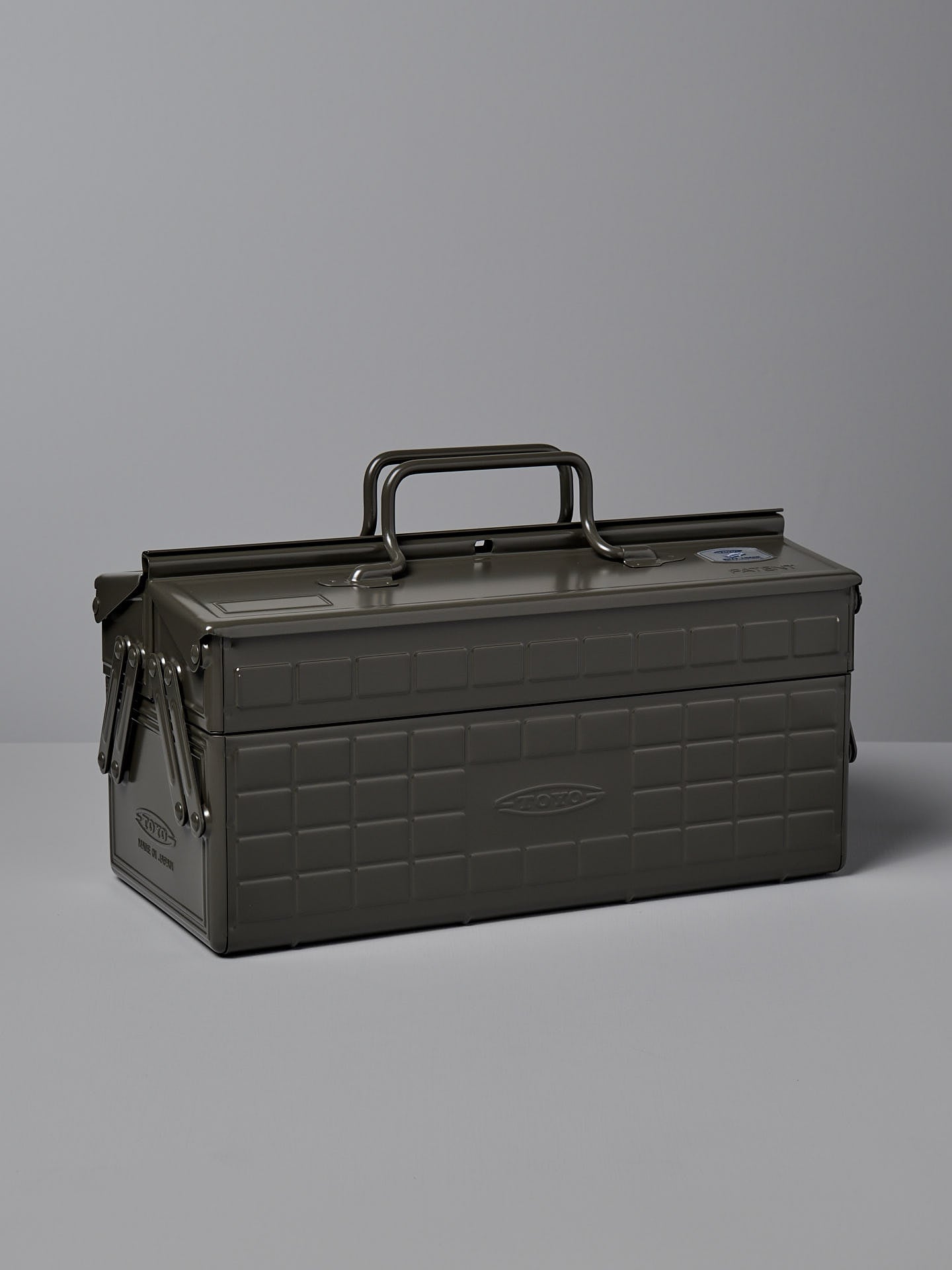 A moss green, powder-coated steel TOYO STEEL Cantilever Toolbox ST-350 with a rectangular shape, clasped lid, and a single handle on top, placed on a gray surface. Made in Osaka.