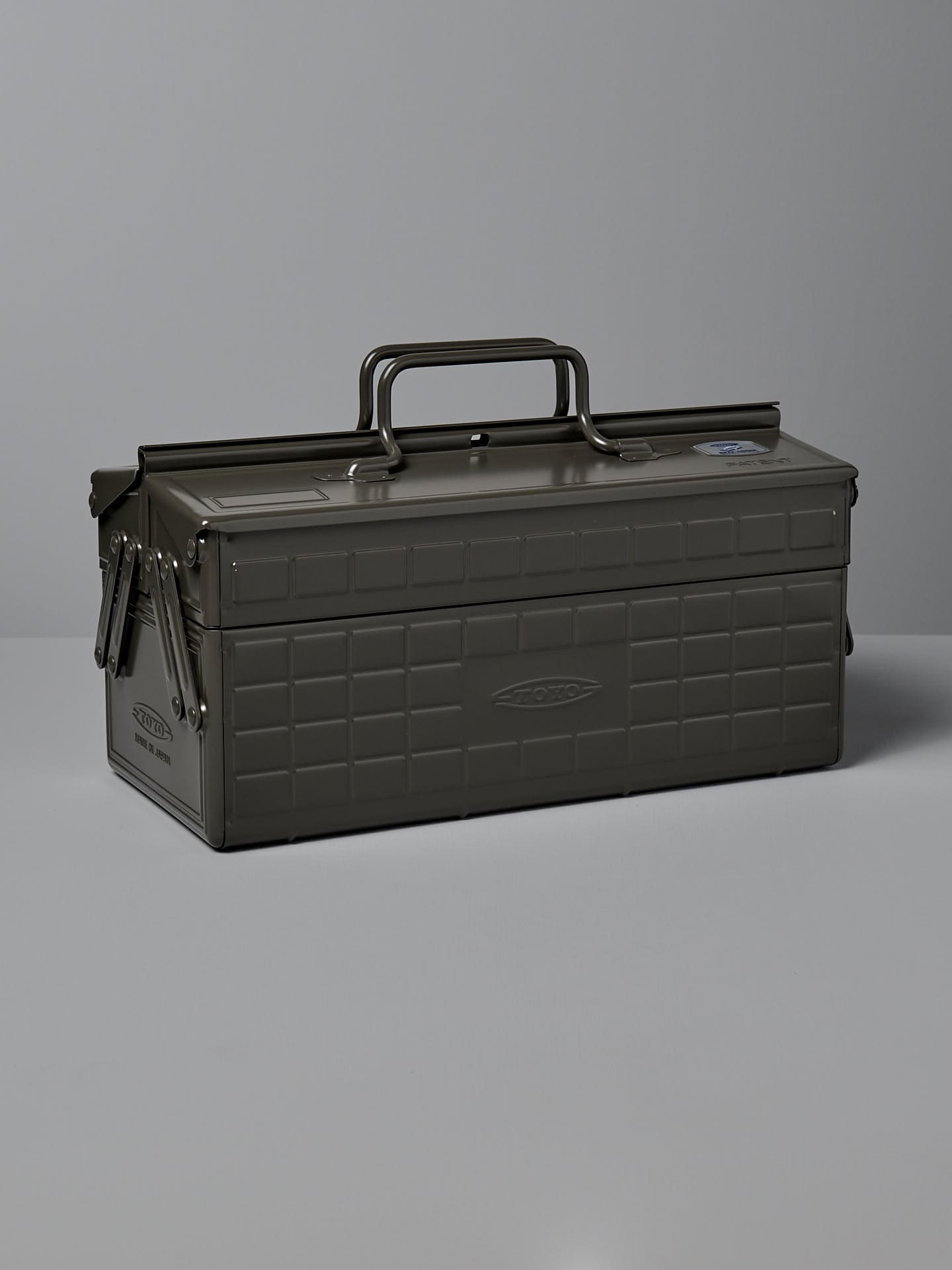 A moss green, powder-coated steel TOYO STEEL Cantilever Toolbox ST-350 with a rectangular shape, clasped lid, and a single handle on top, placed on a gray surface. Made in Osaka.