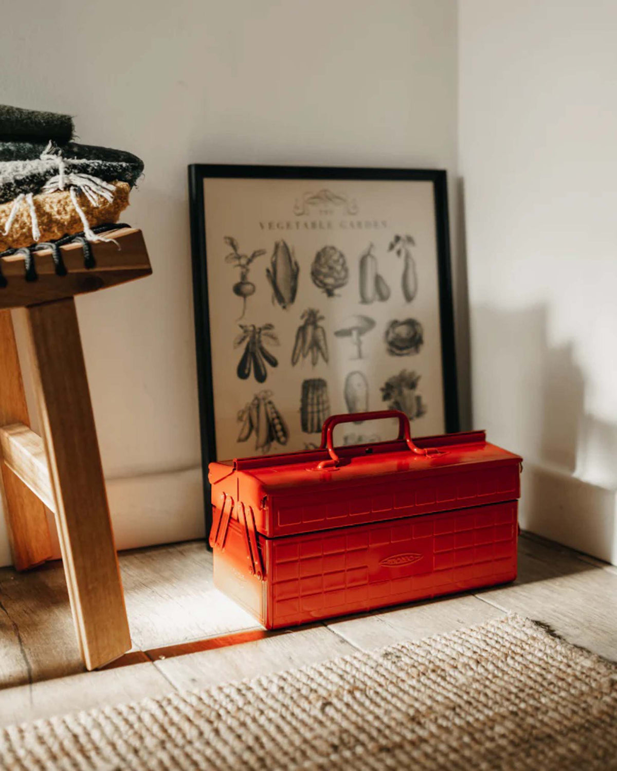 A red metal toolbox on a wooden floor in front of a framed vegetable garden print.