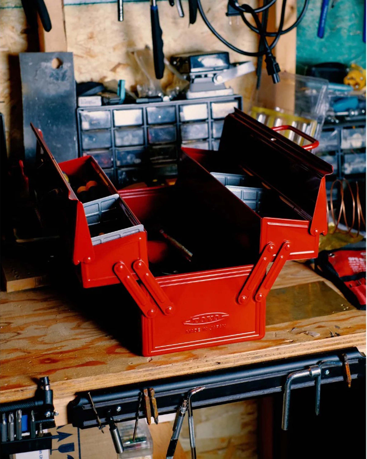 An open red cantilever metal toolbox sitting on a wooden workbench.