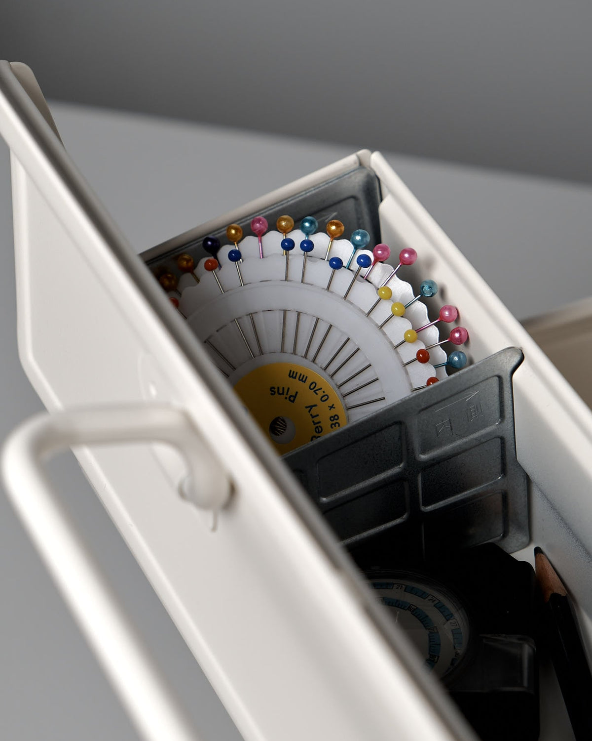 A close-up of a TOYO STEEL Cantilever Toolbox ST-350 – White made from powder-coated steel holds colored sewing pins arranged in a circular holder, with other small sewing supplies visible.