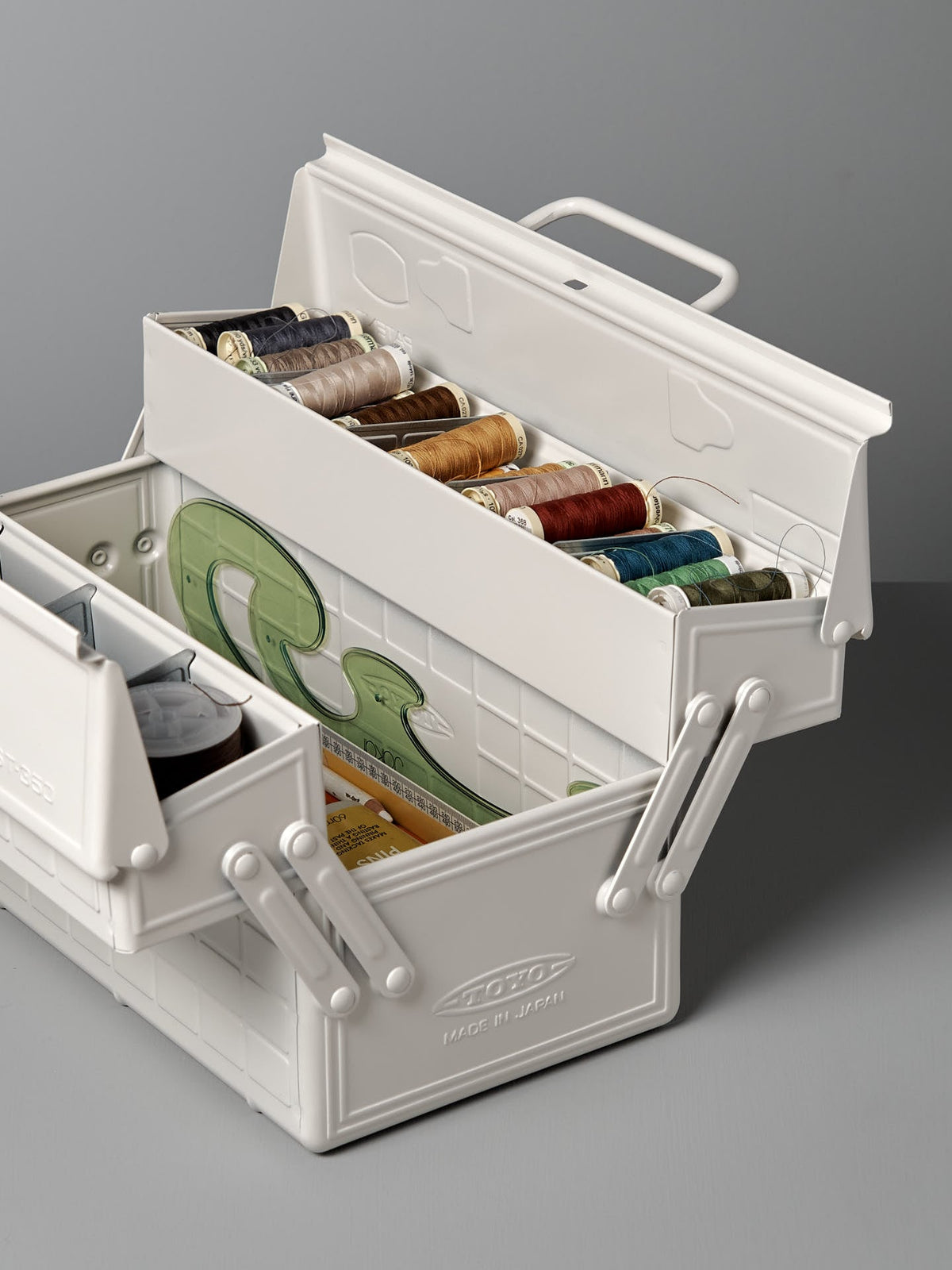 A white powder-coated steel toolbox with multiple compartments contains sewing supplies, including spools of thread in various colors, measuring tape, and other sewing essentials. This Cantilever Toolbox ST-350 – White by TOYO STEEL is a winner of the Good Design Award 2009.