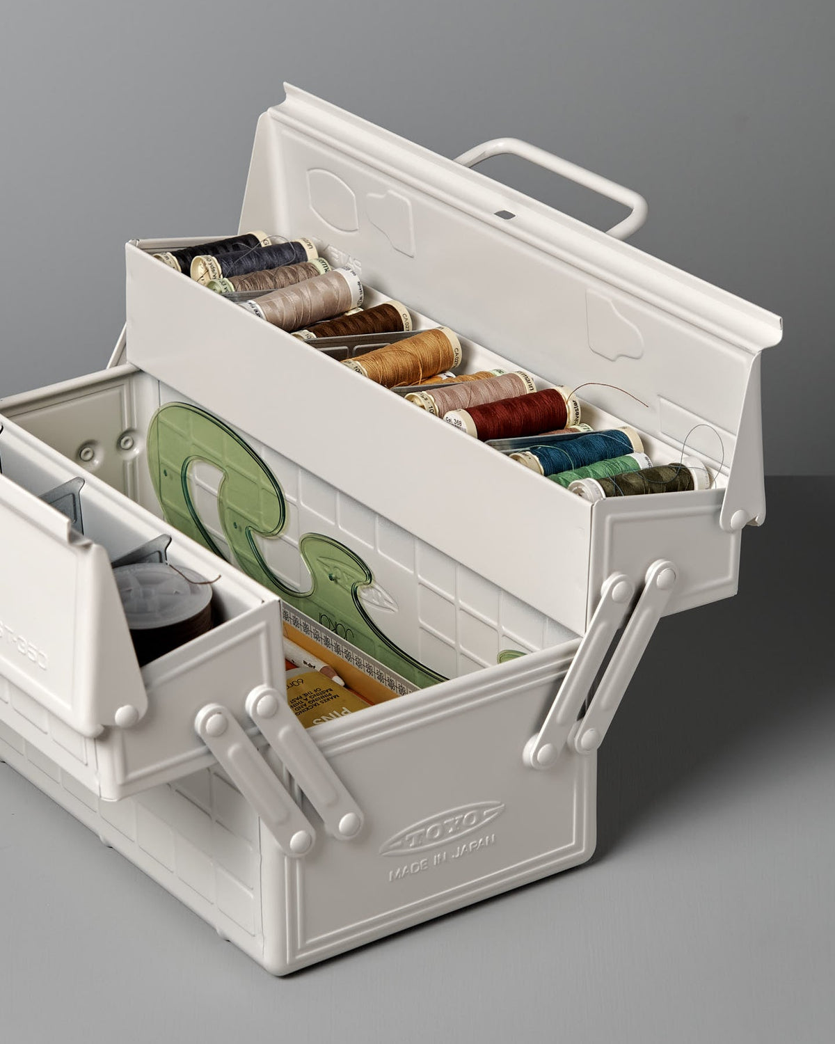 A white powder-coated steel toolbox with multiple compartments contains sewing supplies, including spools of thread in various colors, measuring tape, and other sewing essentials. This Cantilever Toolbox ST-350 – White by TOYO STEEL is a winner of the Good Design Award 2009.