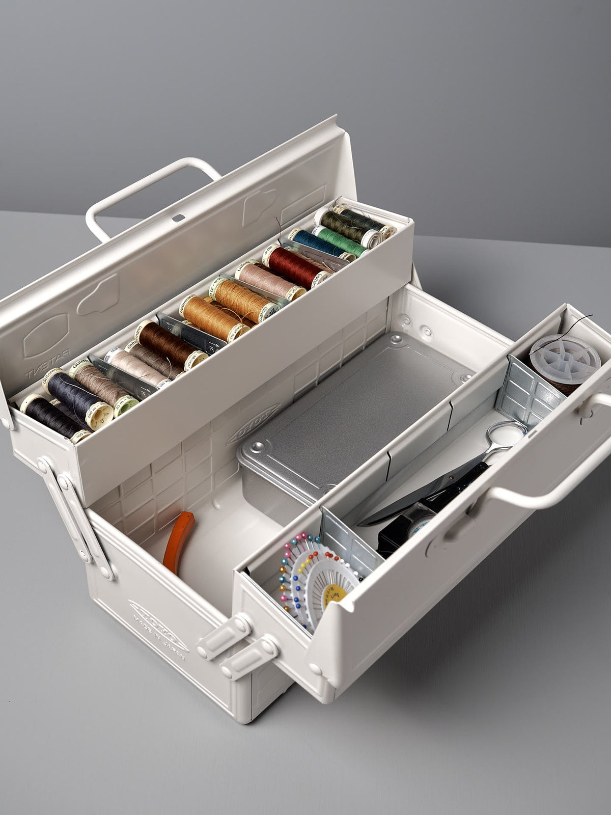 A Cantilever Toolbox ST-350 – White from TOYO STEEL in an iconic camber-top toolbox, crafted from powder-coated steel, reveals various spools of thread, a pair of scissors, a measuring tape, and sewing needles. This design even earned the Good Design Award 2009.