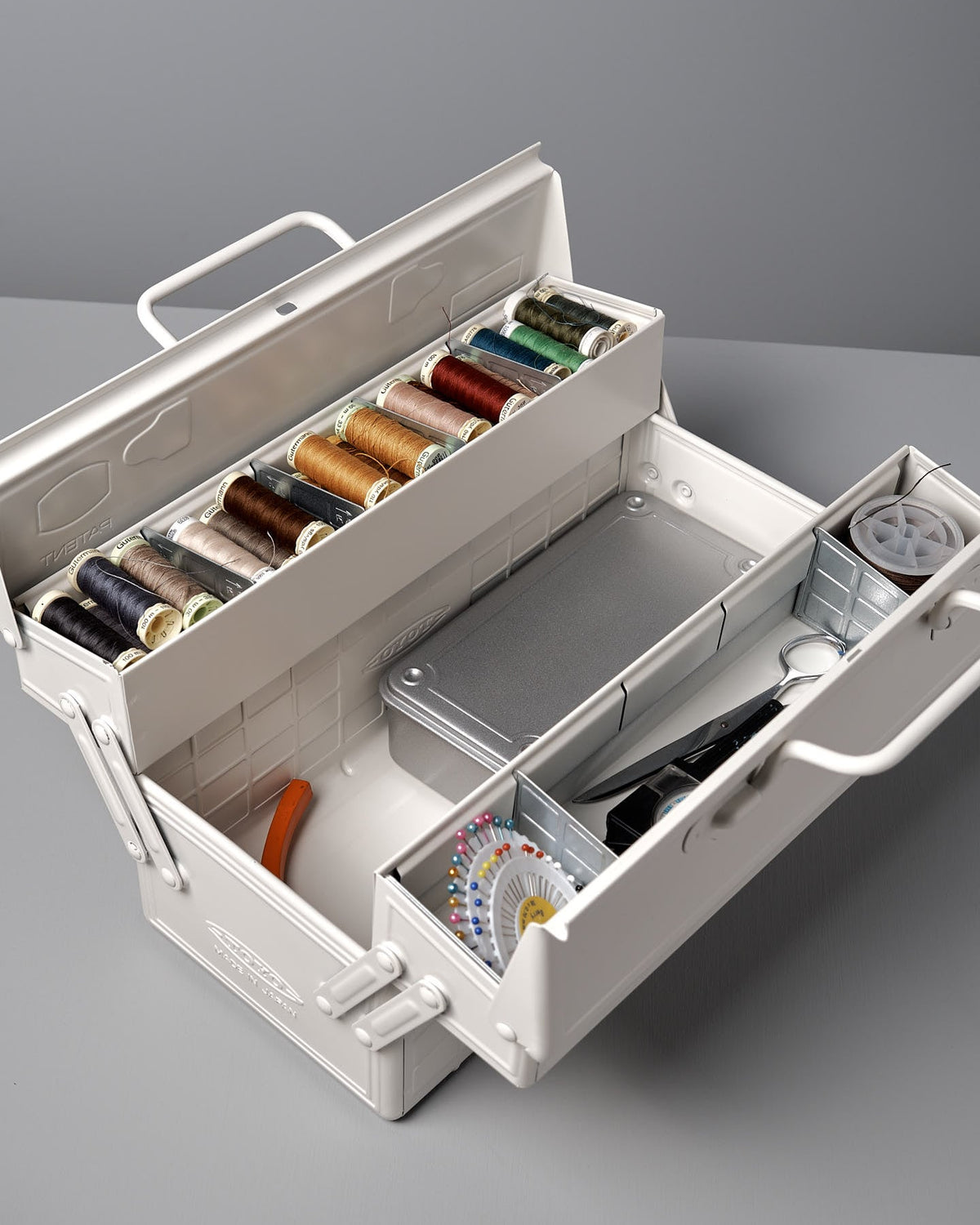 A Cantilever Toolbox ST-350 – White from TOYO STEEL in an iconic camber-top toolbox, crafted from powder-coated steel, reveals various spools of thread, a pair of scissors, a measuring tape, and sewing needles. This design even earned the Good Design Award 2009.