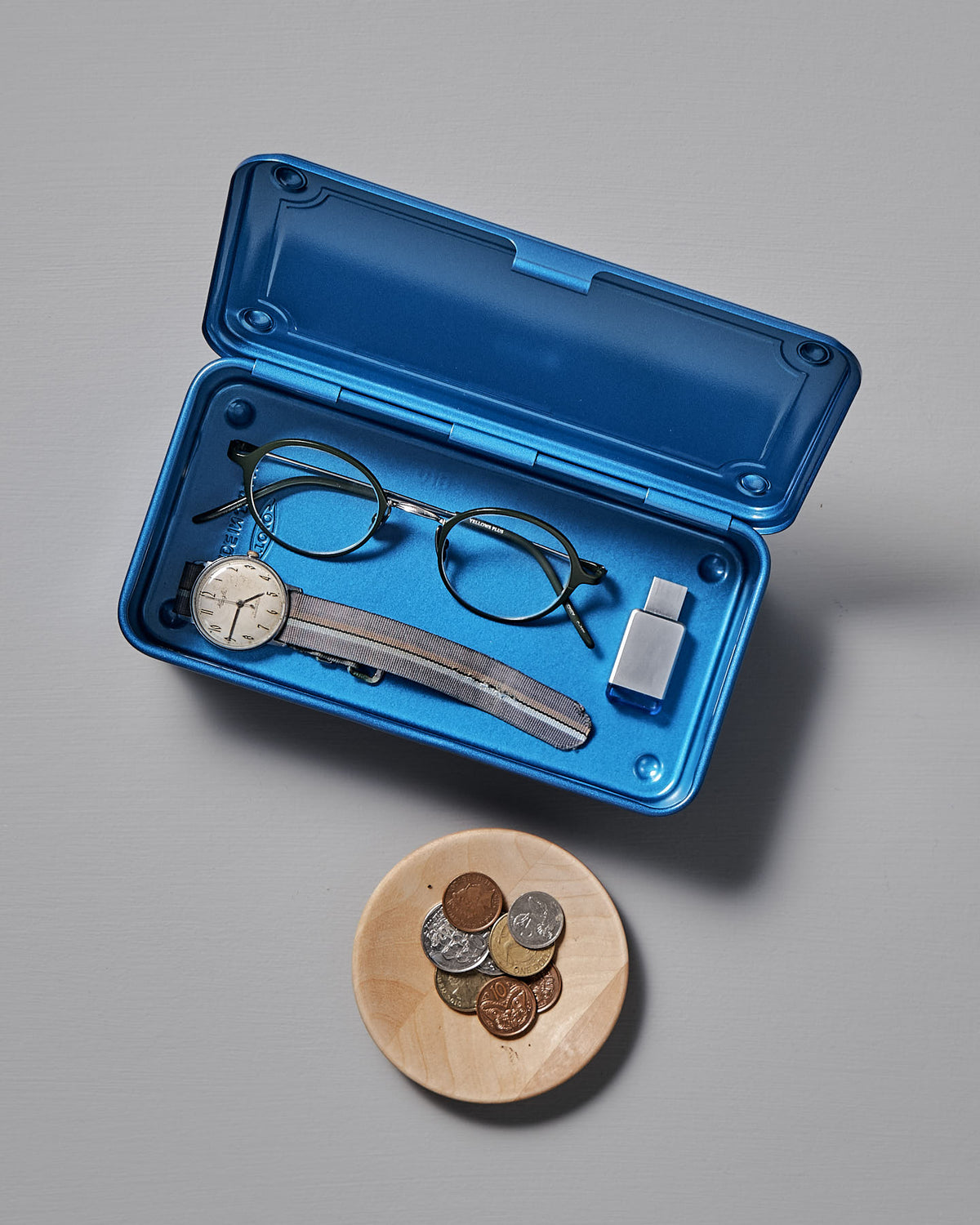 A versatile storage solution is the Mini Steel Toolbox T-190 in blue from TOYO STEEL, containing glasses, a wristwatch, and a USB drive, positioned beside a wooden bowl filled with assorted coins on a gray surface.