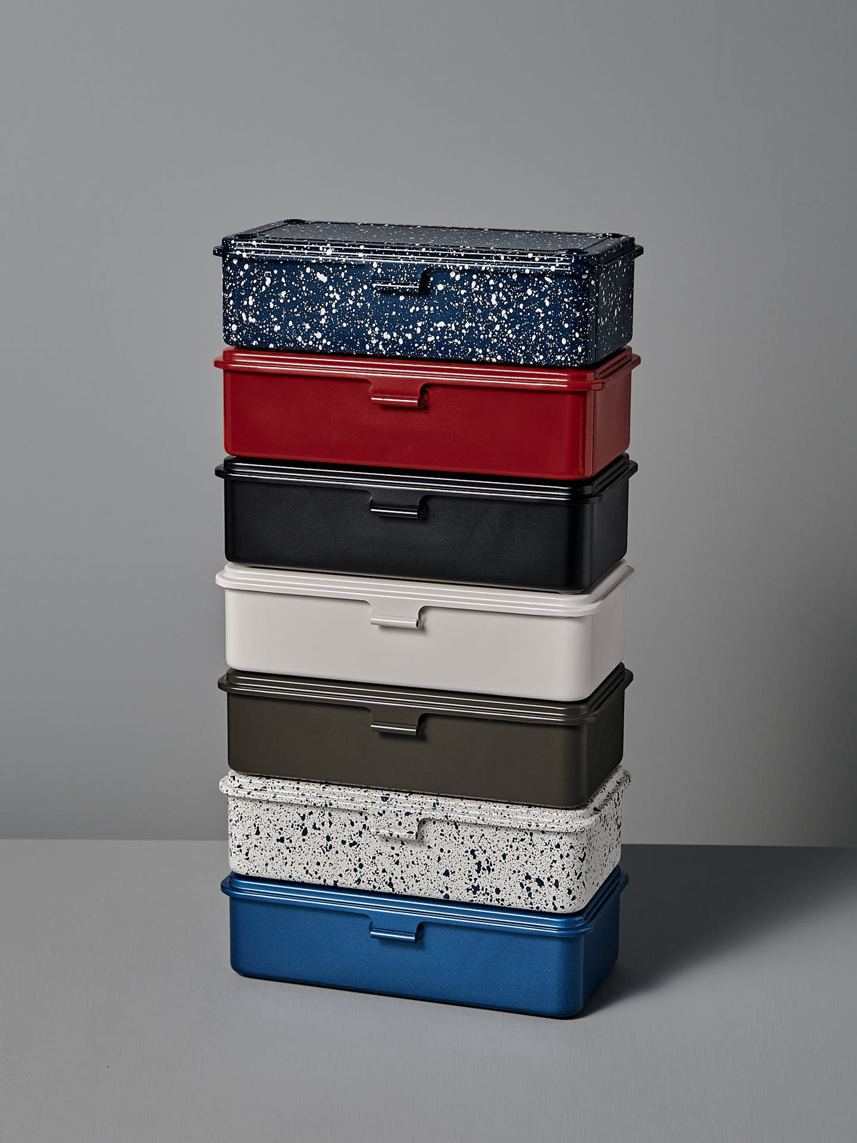 A stack of six versatile storage boxes of different colors: red, black, white, dark green, white with a speckled pattern, and blue (specifically the **Mini Steel Toolbox T-190 – Blue** by **TOYO STEEL**), arranged vertically against a plain grey background. These powder-coated steel storage boxes offer both style and durability.