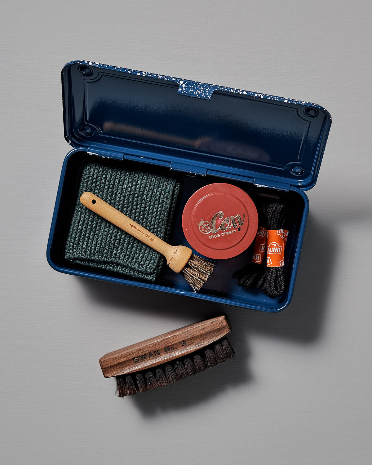 The Mini Steel Toolbox T-190 – Japanese Indigo by TOYO STEEL comes in a sleek, powder-coated steel storage box and includes shoe cream, a small brush, cloth, larger brush, laces, and polish. It is displayed open on a gray surface for easy access and organization.