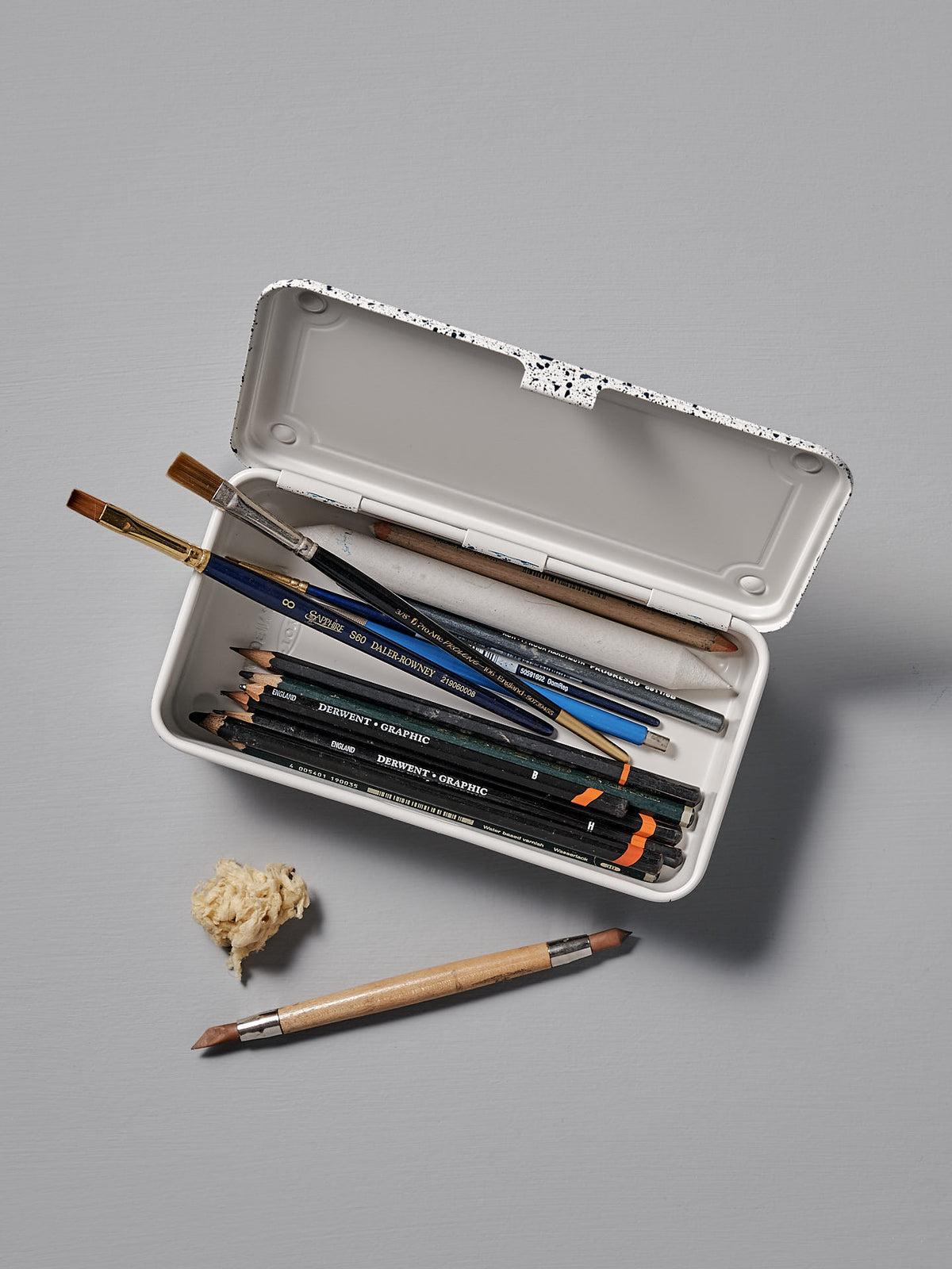 A Mini Steel Toolbox T-190 – Japanese White Indigo by TOYO STEEL, with its versatile design, showcases an array of pencils, brushes, and an eraser on a gray surface. A sharpened pencil and a small pile of sharpener shavings rest outside the toolbox.