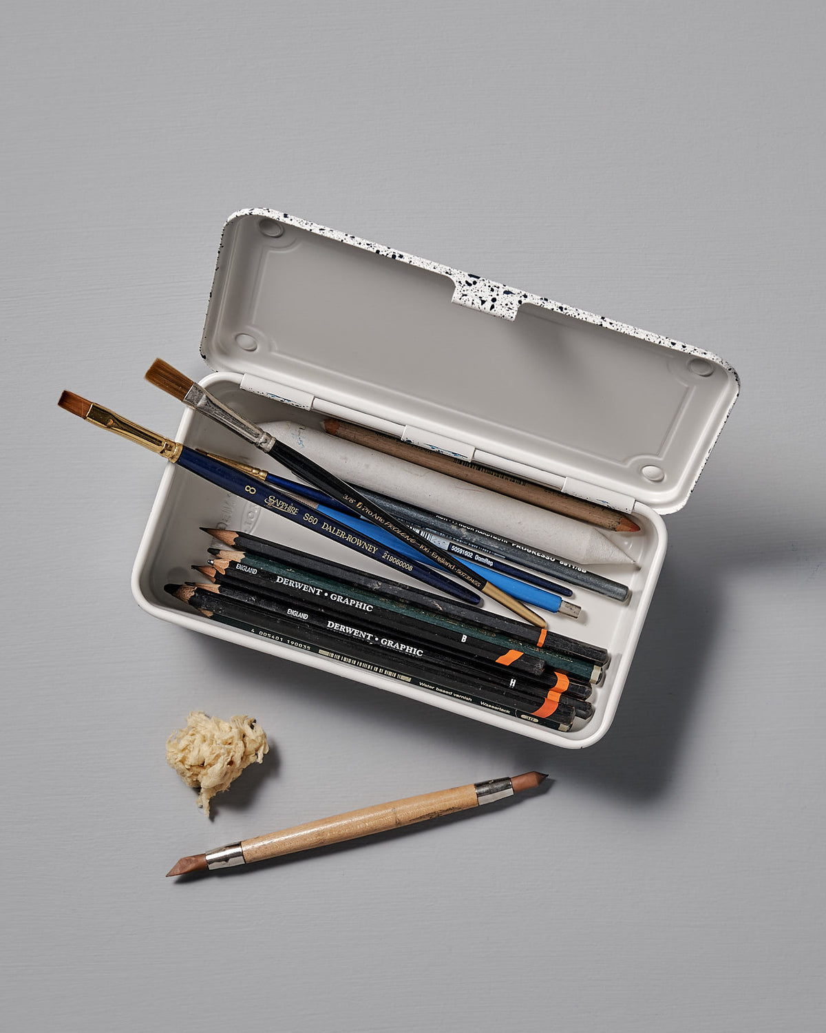 A Mini Steel Toolbox T-190 – Japanese White Indigo by TOYO STEEL, with its versatile design, showcases an array of pencils, brushes, and an eraser on a gray surface. A sharpened pencil and a small pile of sharpener shavings rest outside the toolbox.