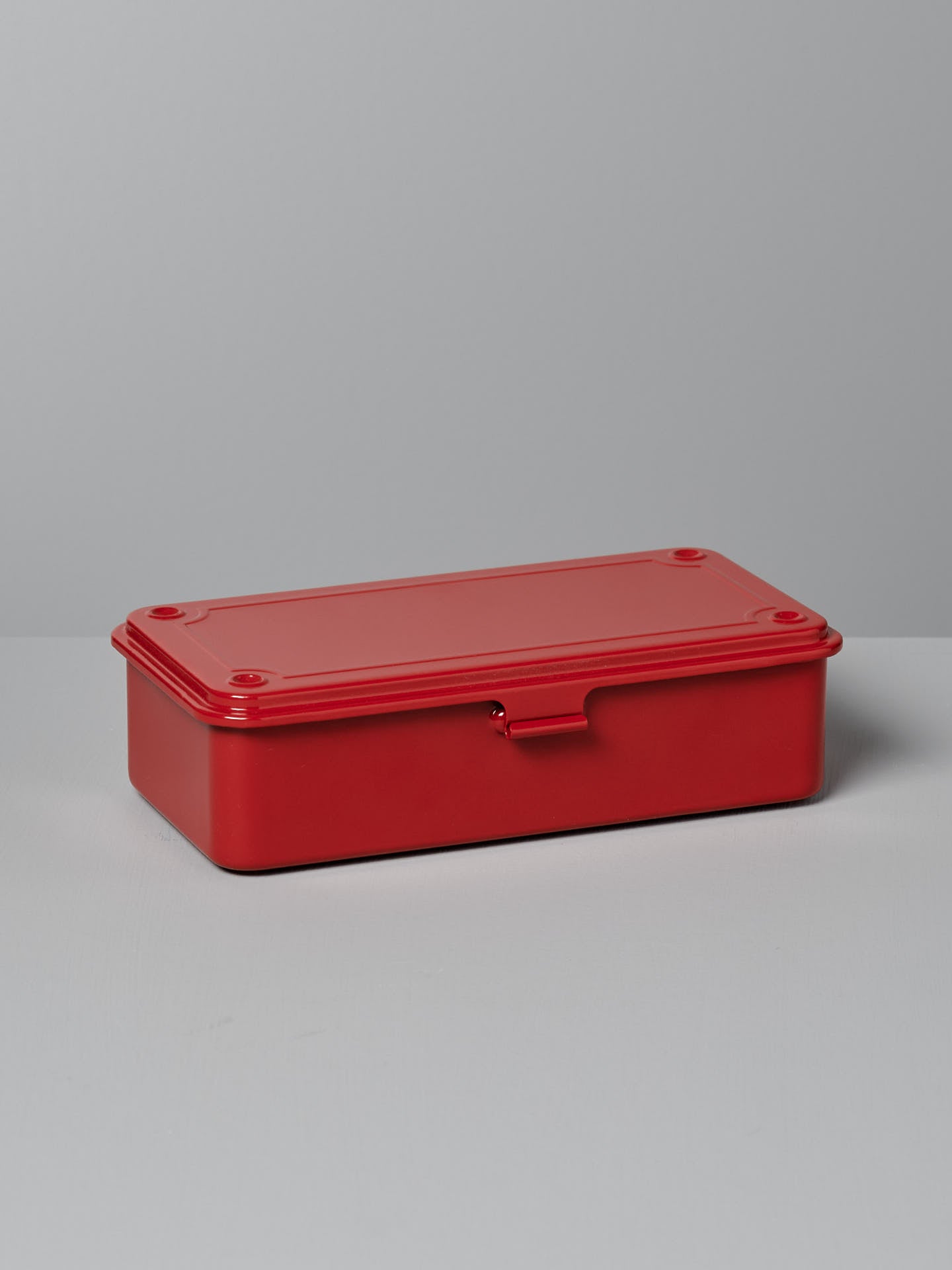 A rectangular, matte red Mini Steel Toolbox T-190 – Red by TOYO STEEL with a snap closure, resting on a gray surface against a light gray background, designed to complement any powder-coated steel aesthetic.