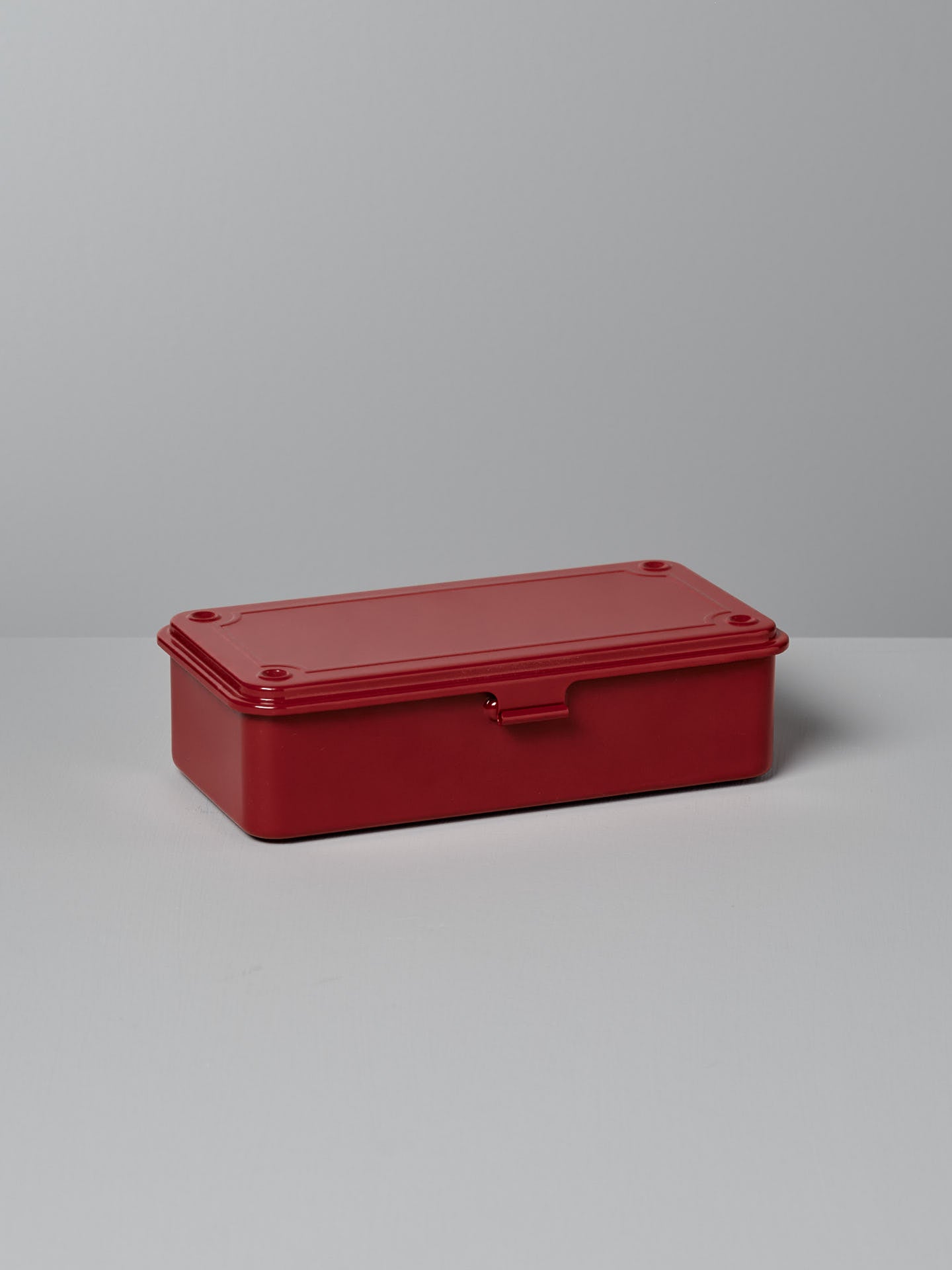 A Mini Steel Toolbox T-190 in red by TOYO STEEL, featuring a seamless rectangular design and a flat lid, sits on a light gray surface.