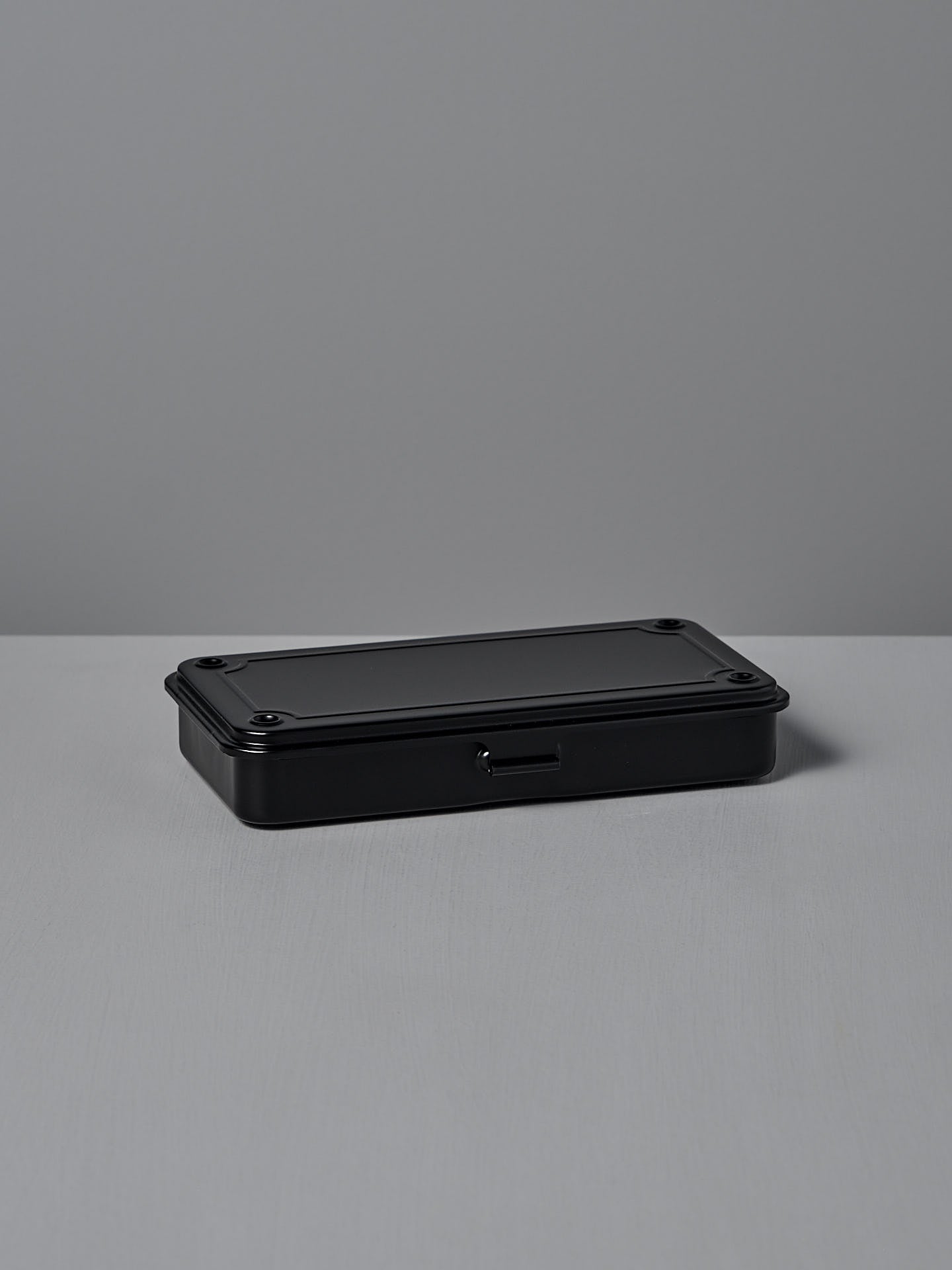 The Mini Steel Toolbox T-192 – Black by TOYO STEEL, crafted from powder-coated steel, is a sleek rectangular tin box with a small latch. It sits elegantly on a light gray surface against a gray backdrop, providing versatile and stylish storage for any space.