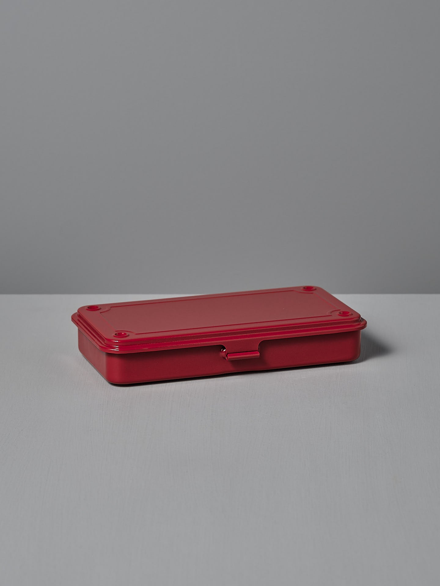 The Mini Steel Toolbox T-192 – Red by TOYO STEEL, featuring a hinged lid and powder-coated steel in red, is elegantly placed on a gray surface with a matching background.