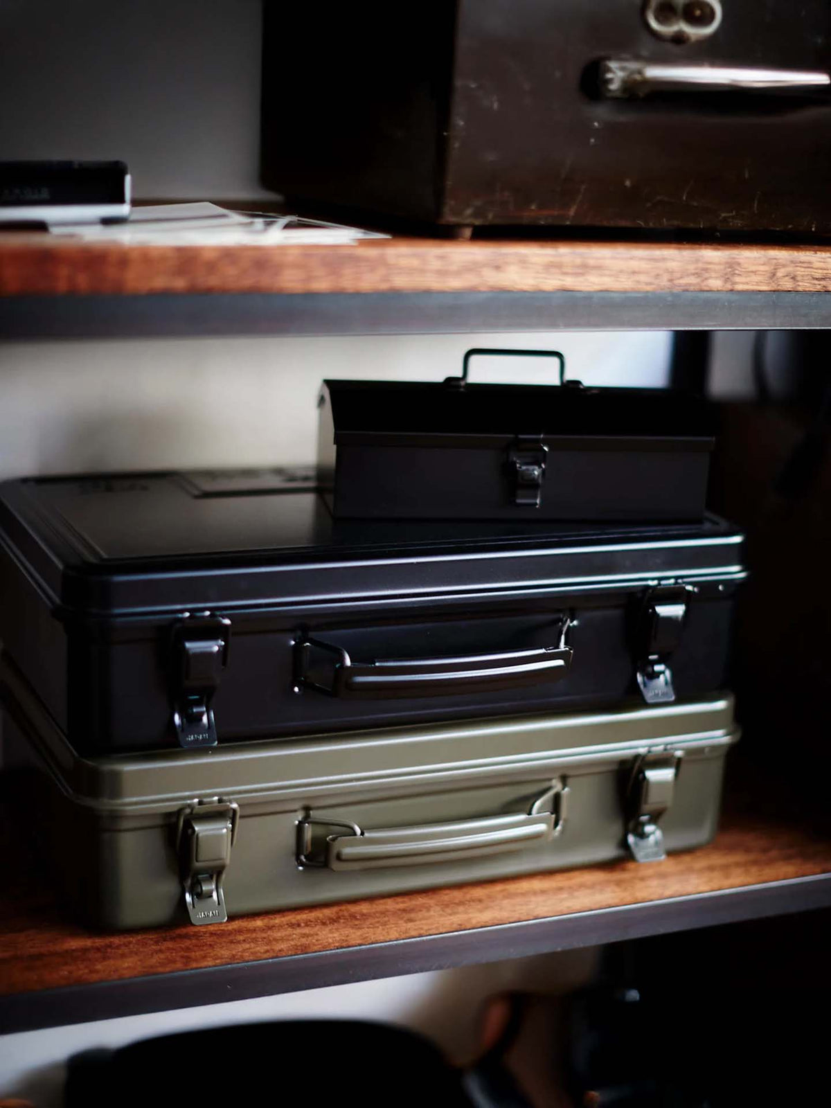 A Trunk-style Toolbox T-360 in moss green is placed atop three Japanese-made steel storage boxes in black and olive green, all stacked on a wooden shelf.