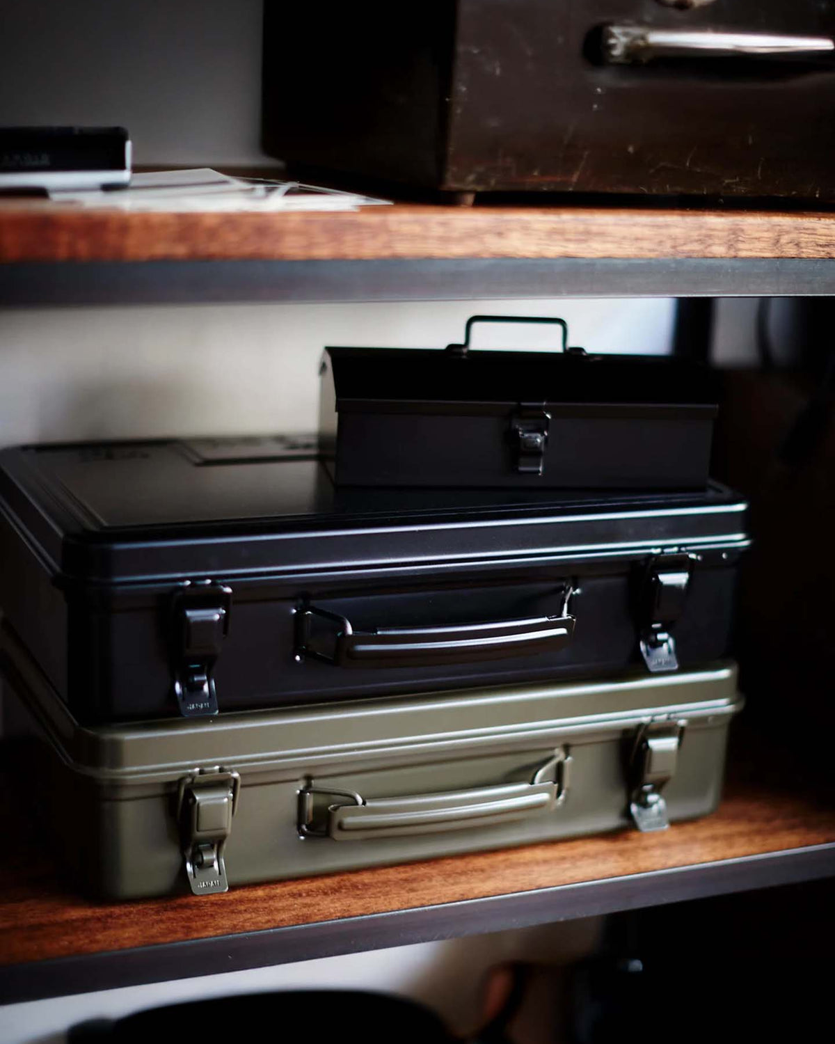 A Trunk-style Toolbox T-360 in moss green is placed atop three Japanese-made steel storage boxes in black and olive green, all stacked on a wooden shelf.