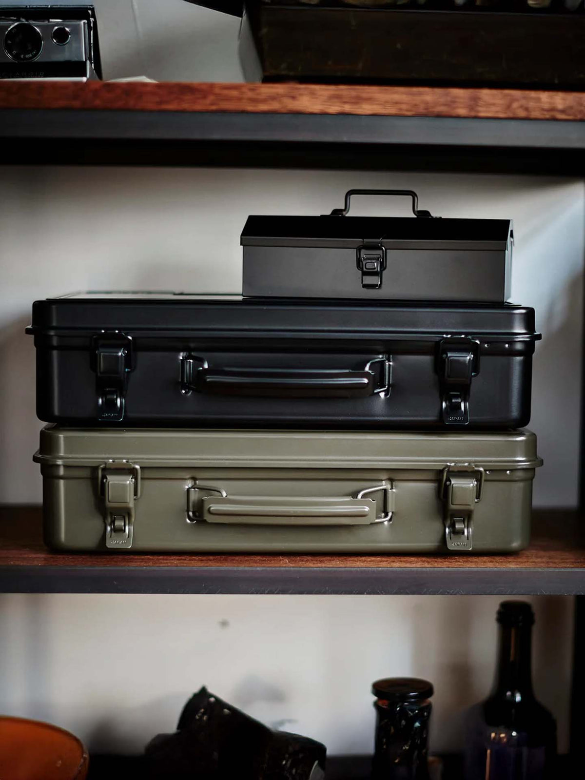 A Trunk-style Toolbox T-360 in Moss Green, made in Japan, is stacked with other steel storage boxes on a wooden shelf amid assorted items.