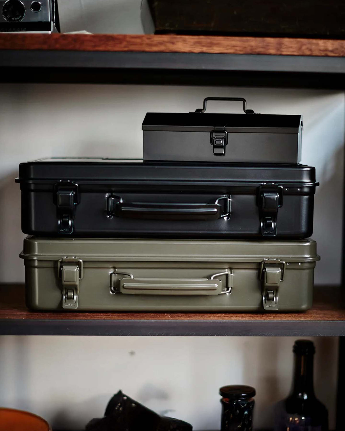 A Trunk-style Toolbox T-360 in Moss Green, made in Japan, is stacked with other steel storage boxes on a wooden shelf amid assorted items.