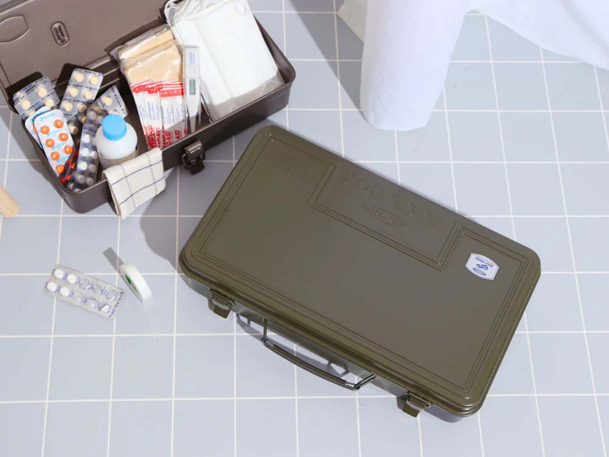 An open first aid kit with medical supplies and medications is shown on a tiled floor next to the Trunk-style Toolbox T-360 in Moss Green, providing a practical storage solution.