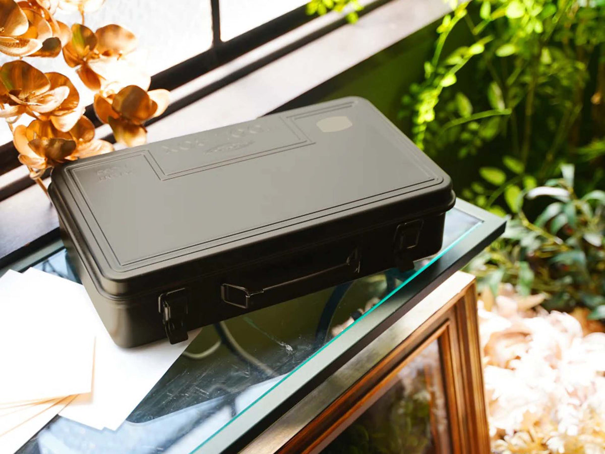 A Moss Green Trunk-style Toolbox T-360 rests on a glass-topped table near a window, surrounded by green plants and papers.
