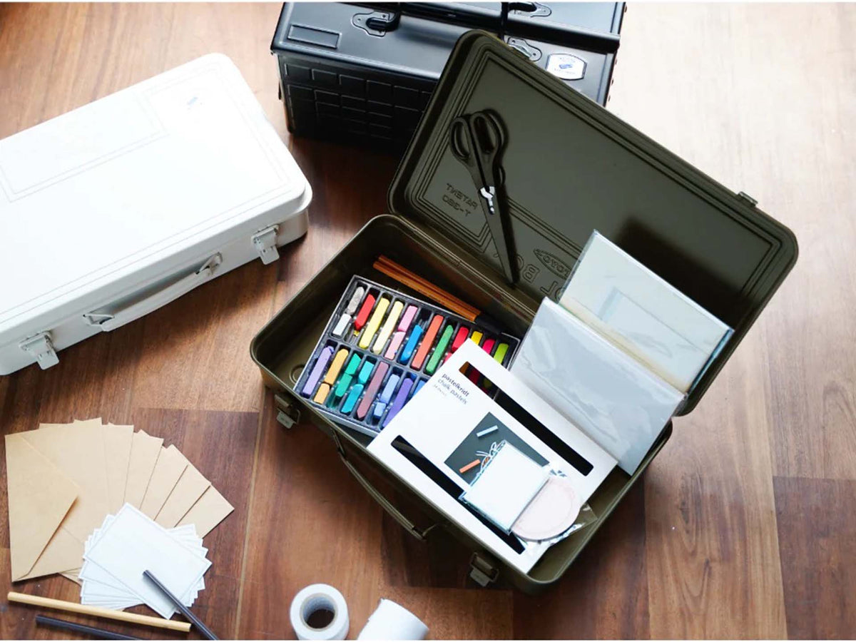 A Trunk-style Toolbox T-360 – Moss Green, made in Japan, holds pastel sticks, scissors, envelopes, and stationery. Two closed cases plus assorted papers and tape are scattered on the wooden floor nearby.