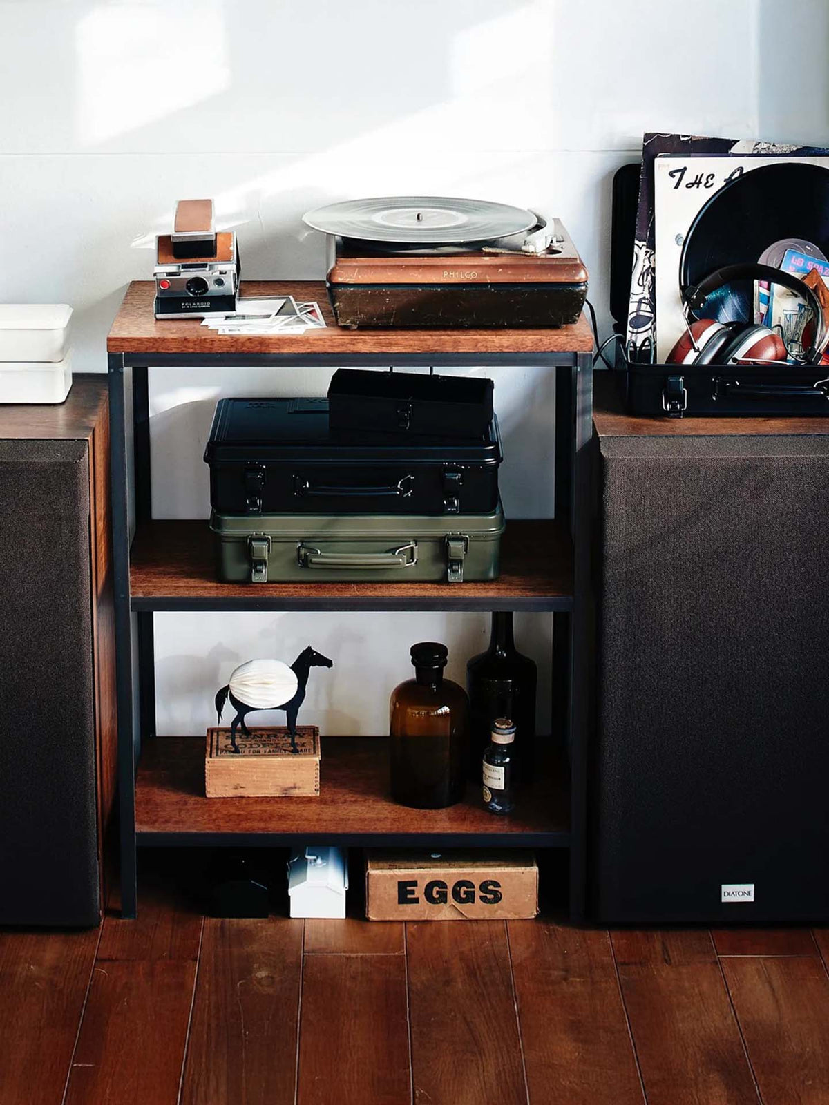 A wooden shelf holds a turntable, suitcases, vinyl records, bottles, a horse figurine, a Trunk-style Toolbox T-360 – Moss Green marked "made in Japan," and an "EGGS" box, flanked by speakers on a wooden floor for eclectic storage.
