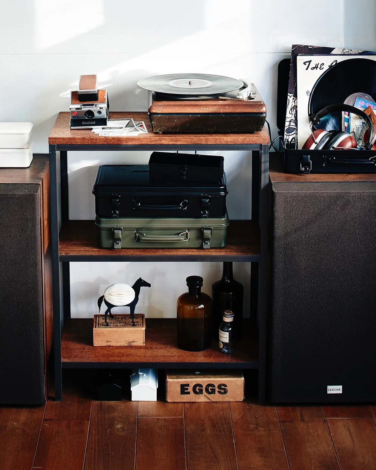 A wooden shelf holds a turntable, suitcases, vinyl records, bottles, a horse figurine, a Trunk-style Toolbox T-360 – Moss Green marked "made in Japan," and an "EGGS" box, flanked by speakers on a wooden floor for eclectic storage.
