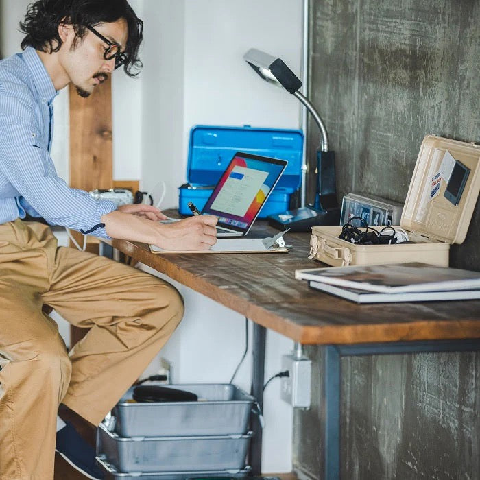 A person dressed in a blue shirt and khaki pants sits at a wooden desk, writing on a notepad. Nearby, a laptop and lamp illuminate the space, while several TOYO STEEL Trunk-style Toolboxes T-360 in white serve as an impeccable storage solution.