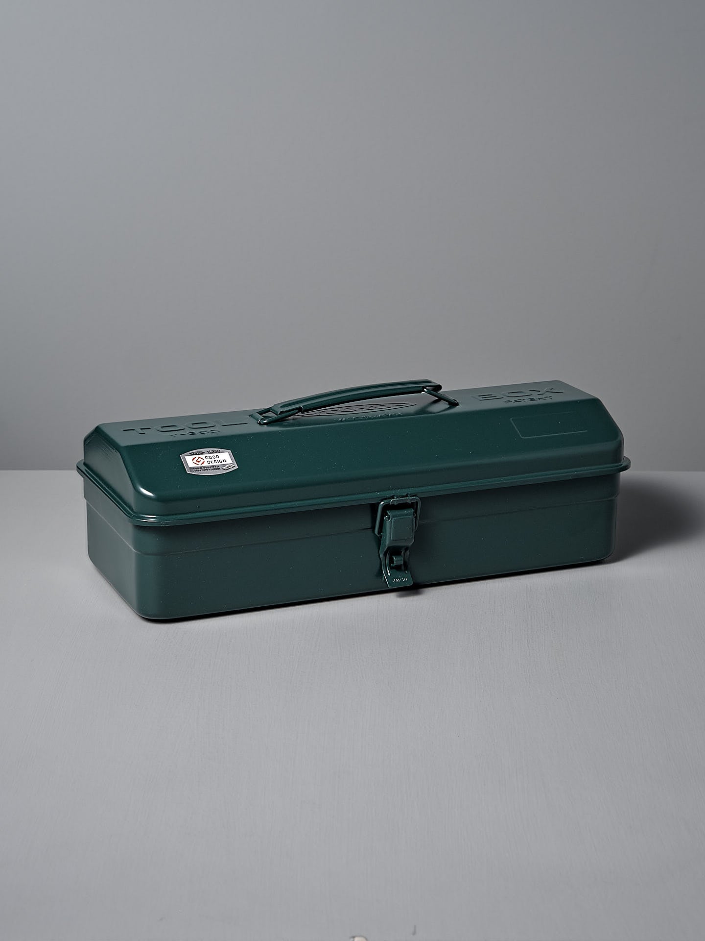 The Camber-Top Toolbox Y-350 by TOYO STEEL, featuring an antique green finish and crafted from powder-coated steel, is elegantly designed with a handle and latch. It rests on a gray surface and earned the Good Design Award in 2009.