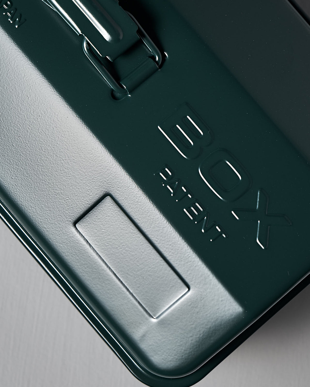 A close-up of the TOYO STEEL Camber-Top Toolbox Y-350 lid in antique green powder-coated steel, embossed with "BOX" and "PATENT," reminiscent of the 2009 Good Design Award style.
