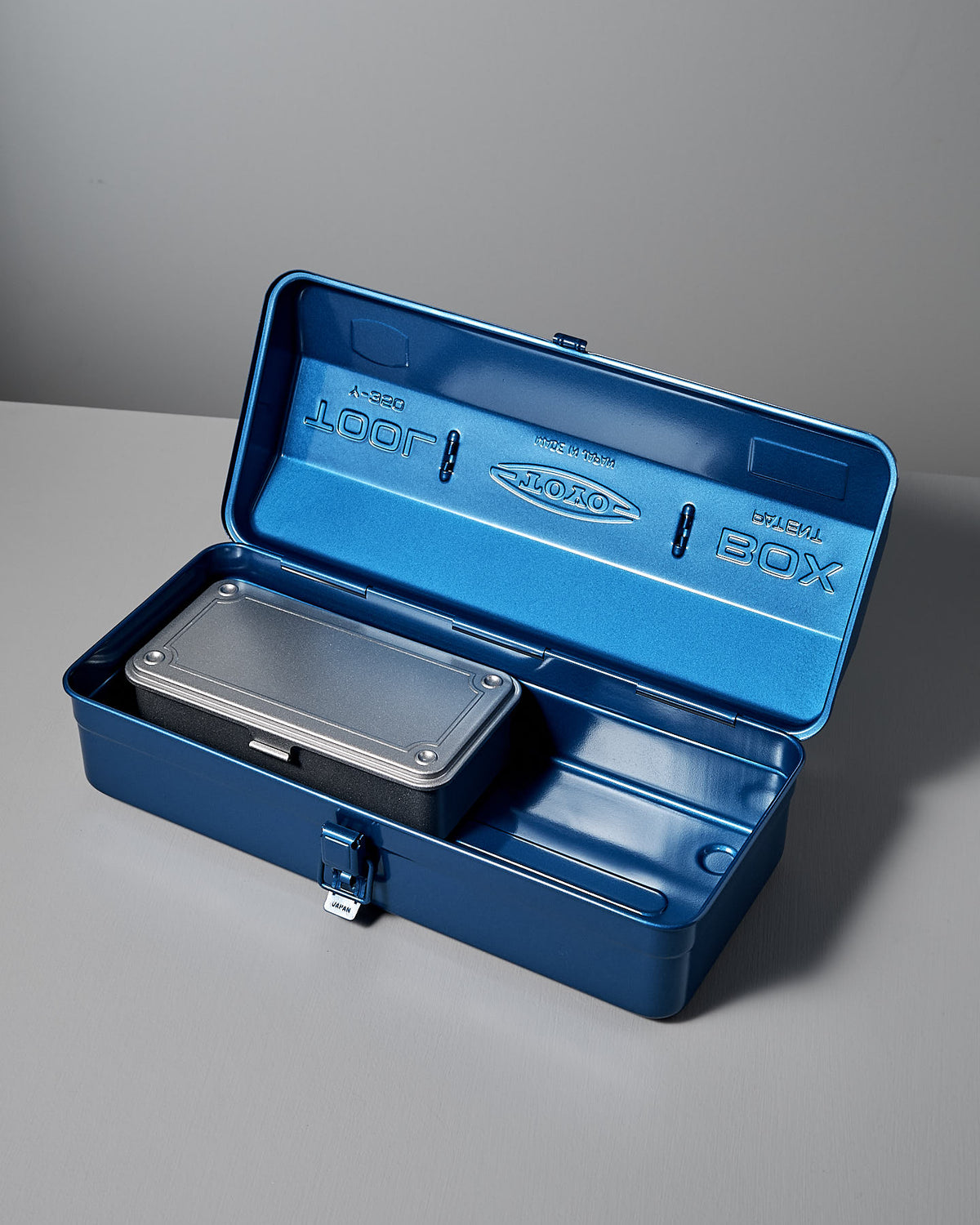 The TOYO STEEL Camber-Top Toolbox Y-350 in blue, constructed from powder-coated steel, elegantly holds a smaller closed container inside as it rests on a gray surface.
