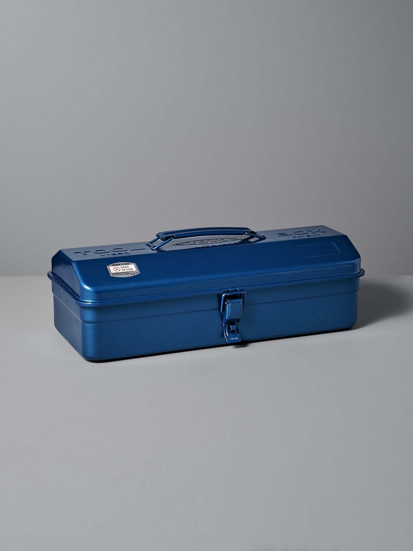 The TOYO STEEL Camber-Top Toolbox Y-350 in blue, featuring a powder-coated steel finish and a sturdy handle with a latch, is elegantly showcased on a gray surface.