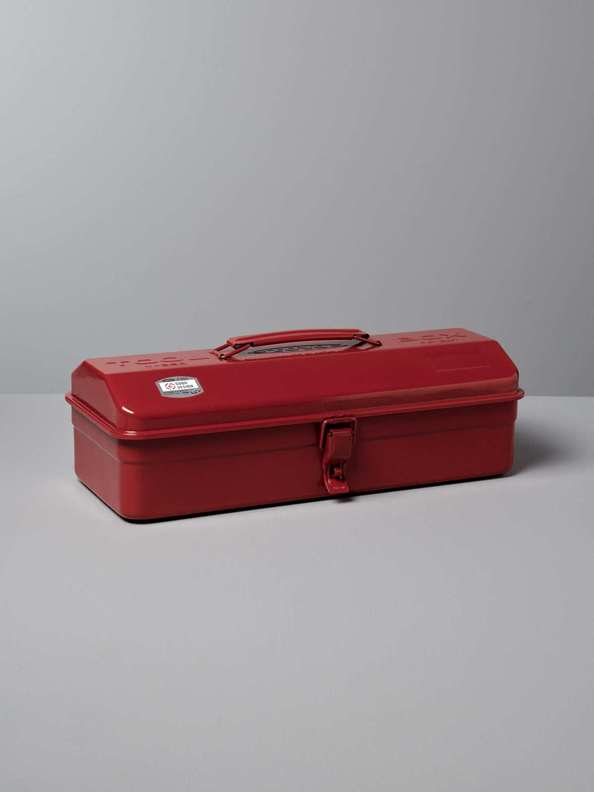 A closed red steel toolbox with a handle on top and a latch on the front, sitting on a gray surface against a gray background — an example of practical design that could easily merit a Good Design Award. This is the TOYO STEEL Camber-Top Toolbox Y-350 – Red.