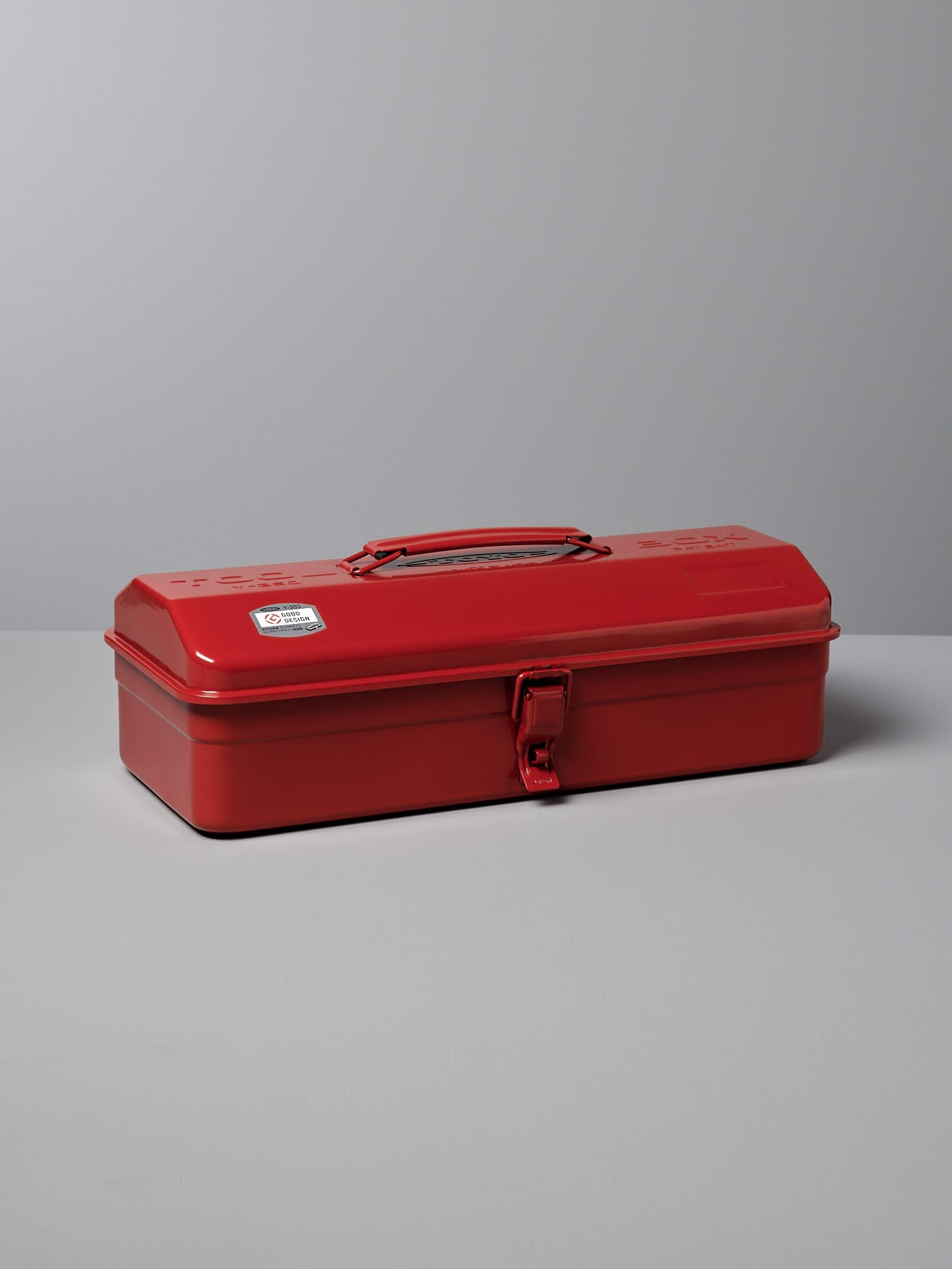 A closed red steel toolbox with a handle on top and a latch on the front, sitting on a gray surface against a gray background — an example of practical design that could easily merit a Good Design Award. This is the TOYO STEEL Camber-Top Toolbox Y-350 – Red.