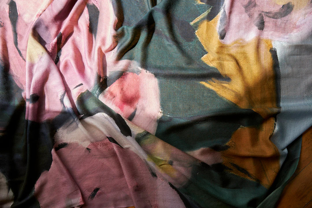 A close-up of an Edith Wool Scarf with an abstract floral pattern in shades of pink, green, yellow, and black, draped and folded. This exquisite piece is hand-painted in oil on 100% wool by Walker &amp; Bing.