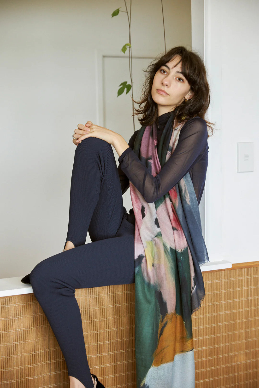 Woman in dark clothing sits on a counter with one knee raised, wearing a Walker &amp; Bing Edith Wool Scarf, and looking at the camera.