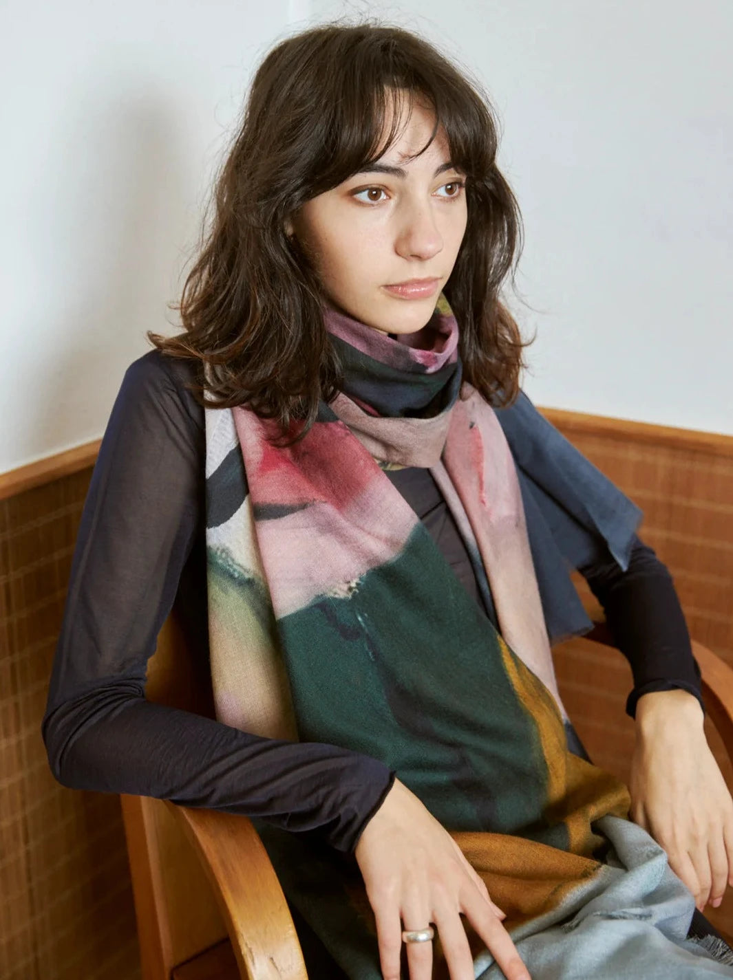 A person with shoulder-length brown hair sits in a wooden chair, wearing a long, Walker & Bing Edith Wool Scarf made of 100% wool over a dark top.