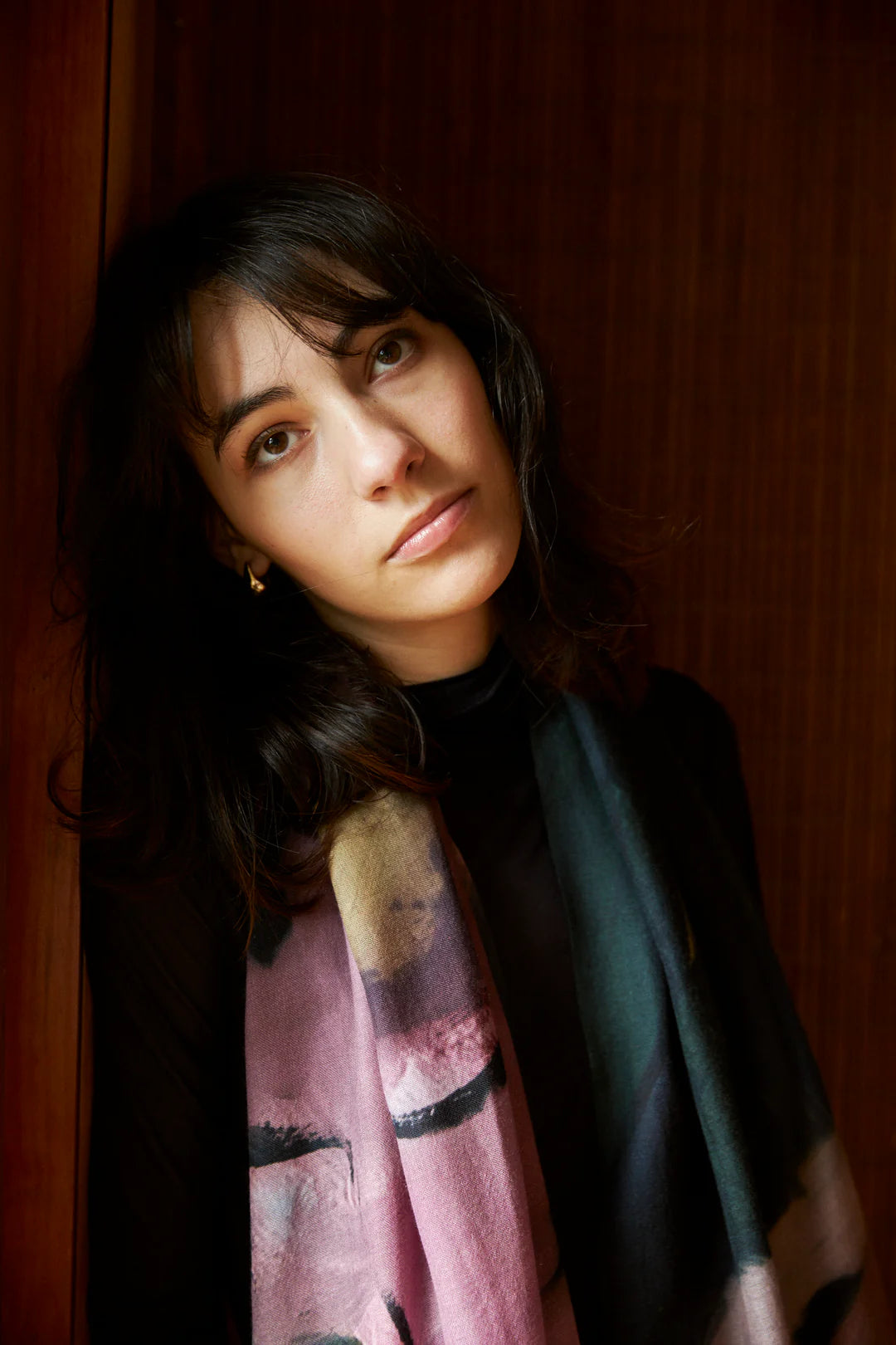 A person with medium-length dark hair and a neutral expression is wearing an Edith Wool Scarf, made from 100% wool by Walker &amp; Bing, and standing against a wooden background.