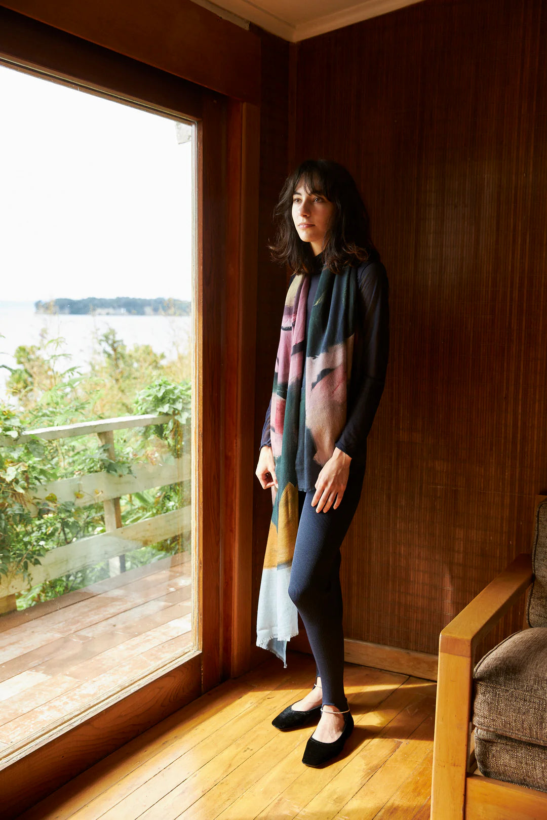 A woman stands indoors near a large window, wearing an Edith Wool Scarf by Walker &amp; Bing, with a dark outfit and black shoes. The scene captures her against a wooden wall, with an outdoor view of greenery and water in the background.