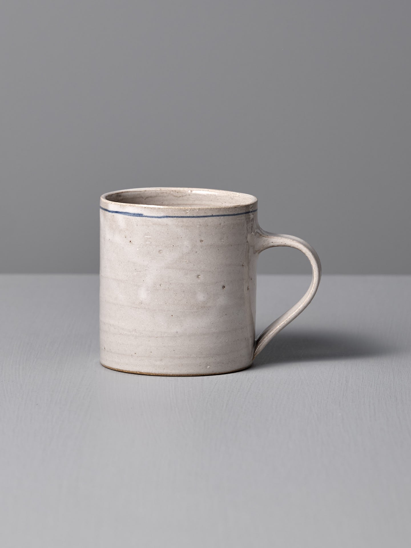 The Zoë Isaacs Coffee Cup – Cobalt Line is a simple, elegant ceramic mug in light gray with a handle and a matte blue-grey glaze, displayed on a matching surface against a gray background.