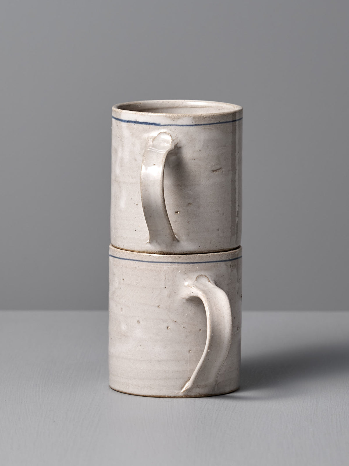 Two Zoë Isaacs Coffee Cup – Cobalt Line ceramic mugs with handles, featuring a matte blue-grey glaze, are stacked vertically on a gray surface against a gray background.