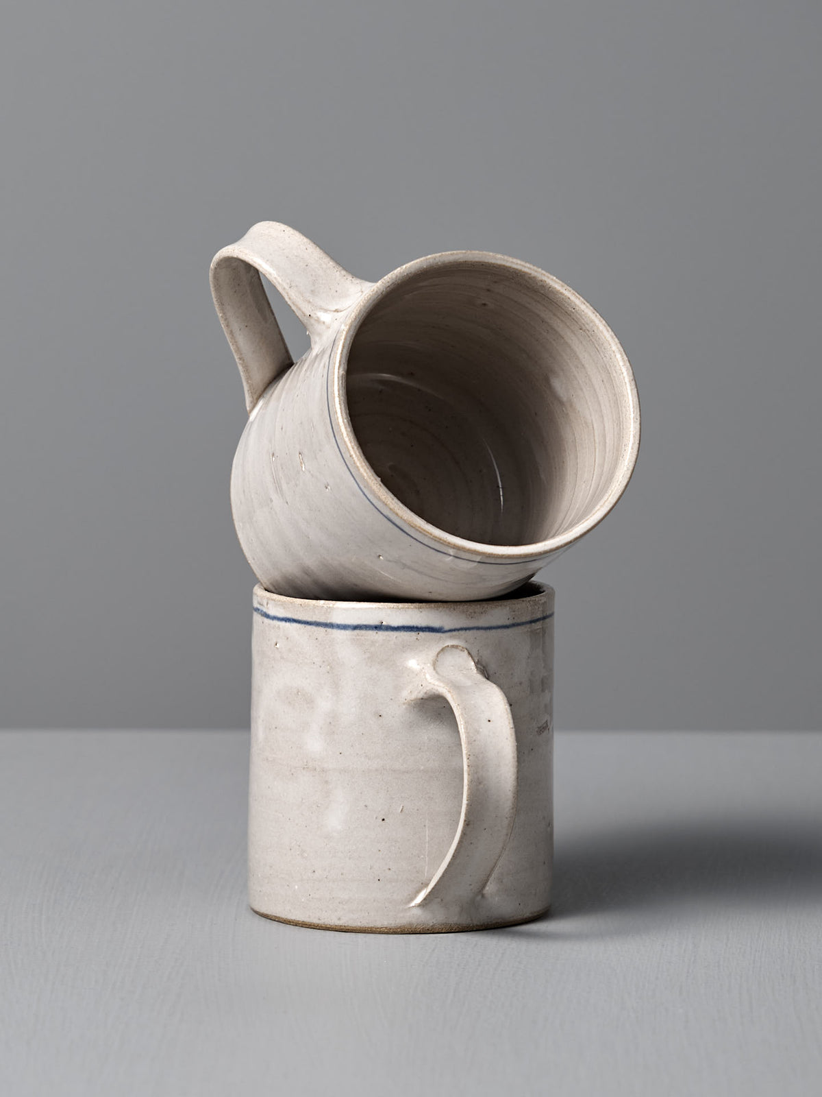 Two Zoë Isaacs Coffee Cup – Cobalt Line mugs: one upright, the other—decorated with a hand-painted matte blue-grey glaze—rests horizontally on top, handle touching the lower mug. Both showcase unique handmade craftsmanship.