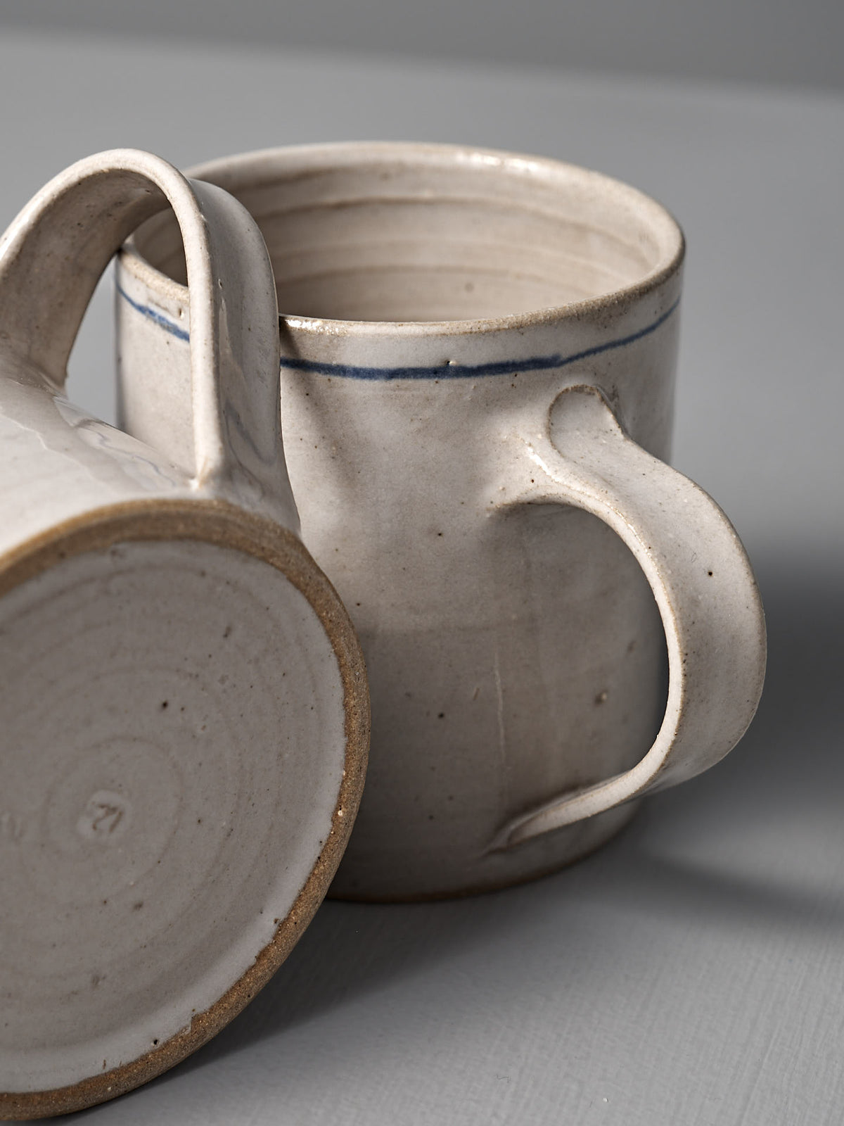 Two Zoë Isaacs Coffee Cup – Cobalt Line mugs: one upright, one on its side, displaying a matte blue-grey glaze with a hand-painted cobalt line near the rim.
