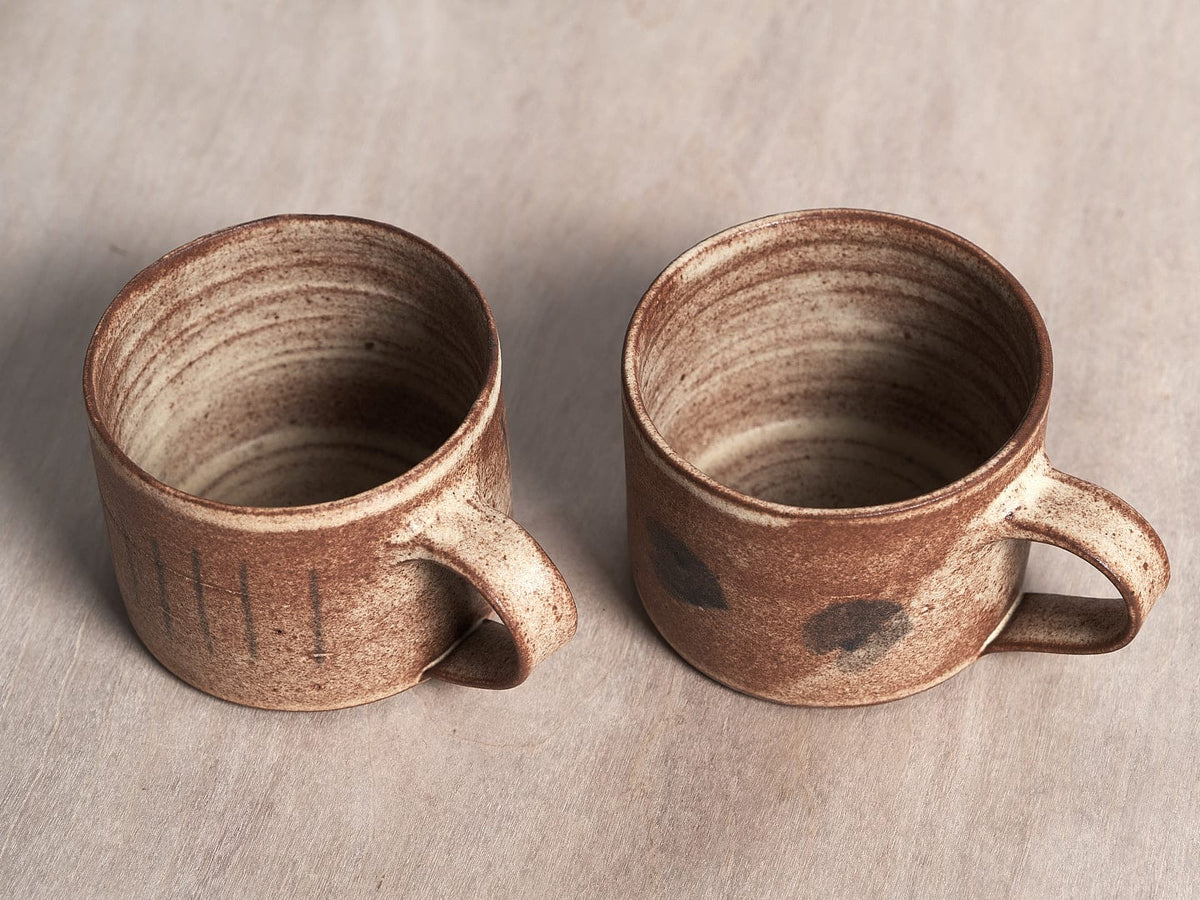 Two handmade brown Spots mugs with a matte cream glaze on a wooden table.