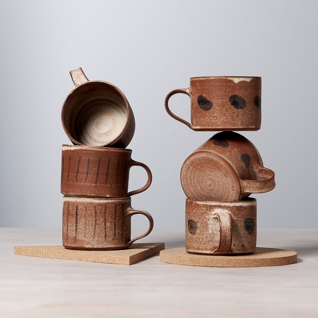 A stack of Zoë Isaacs Stripes mugs made from iron-rich clay, featuring a matte cream glaze and are dishwasher safe, placed on top of a wooden board.