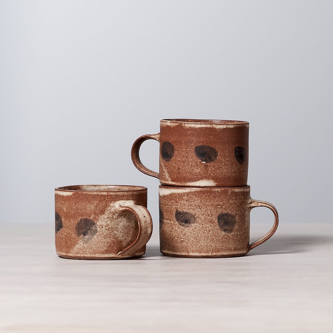 Three brown and black Zoë Isaacs Spots mugs, made from iron-rich clay, with a matte cream glaze finish, sitting on top of a wooden table.