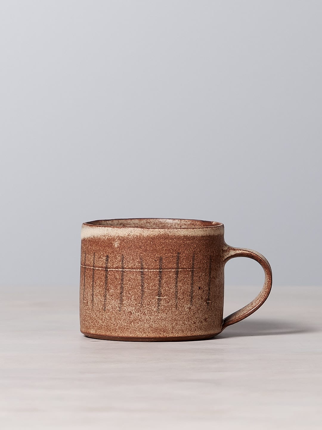 A small brown Zoë Isaacs mug made of iron-rich clay, with a matte cream glaze. This dishwasher safe Stripes mug is sitting on a table.