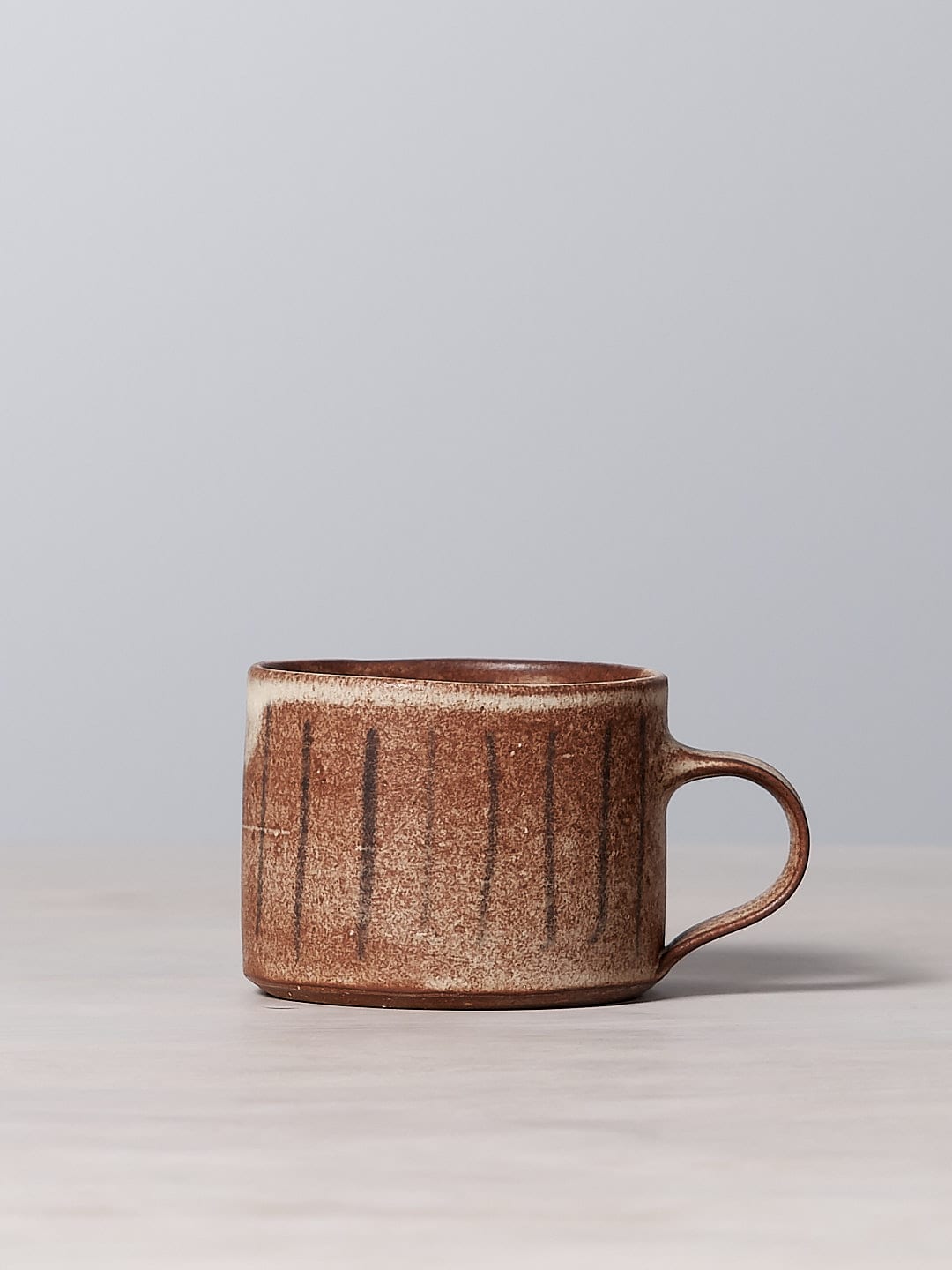A small brown Mug – Stripes made from iron-rich clay, featuring a dishwasher safe design and a matte cream glaze, sitting on a table.