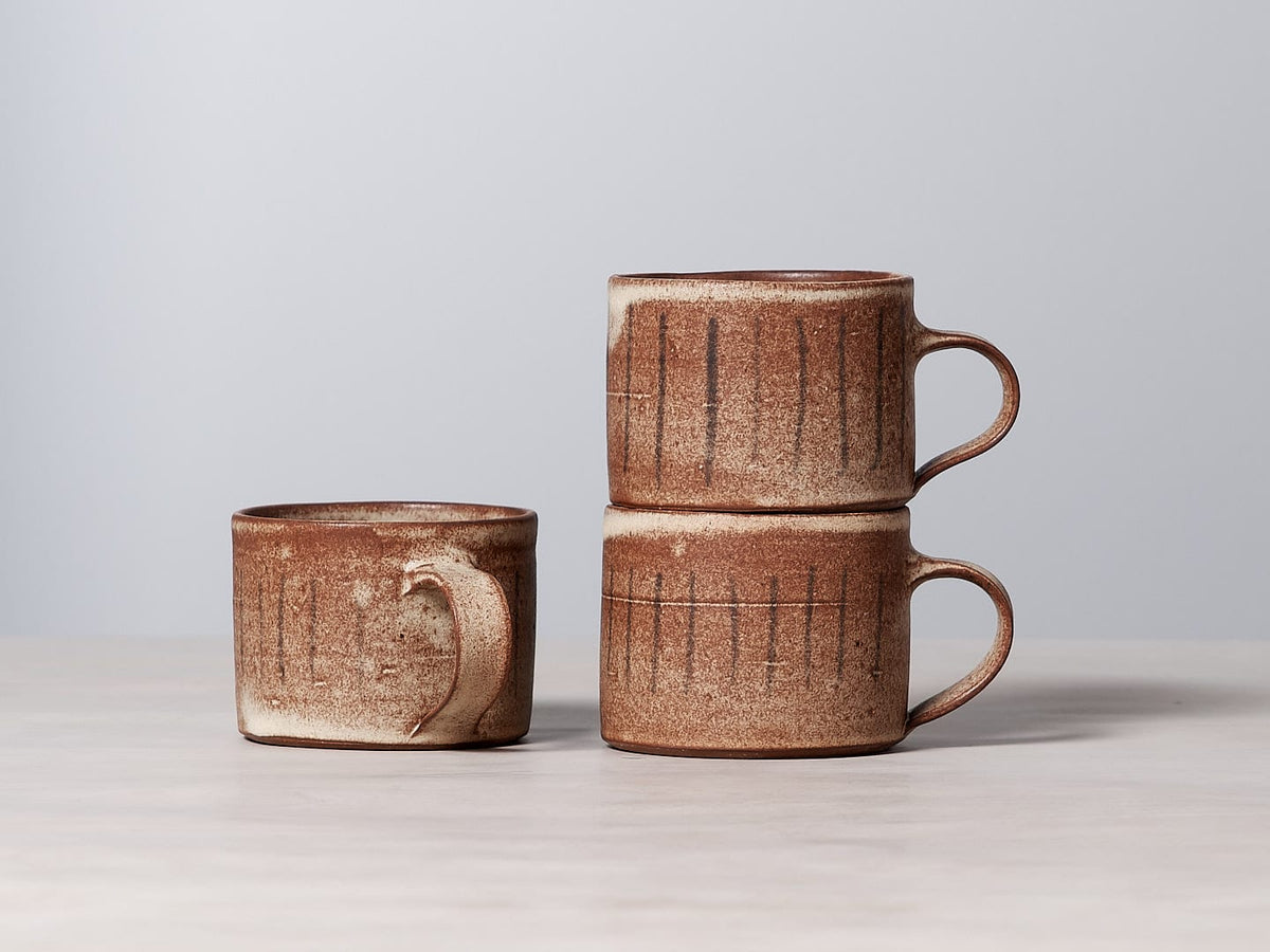 Three brown Stripes mugs with a matte cream glaze sitting on a table.