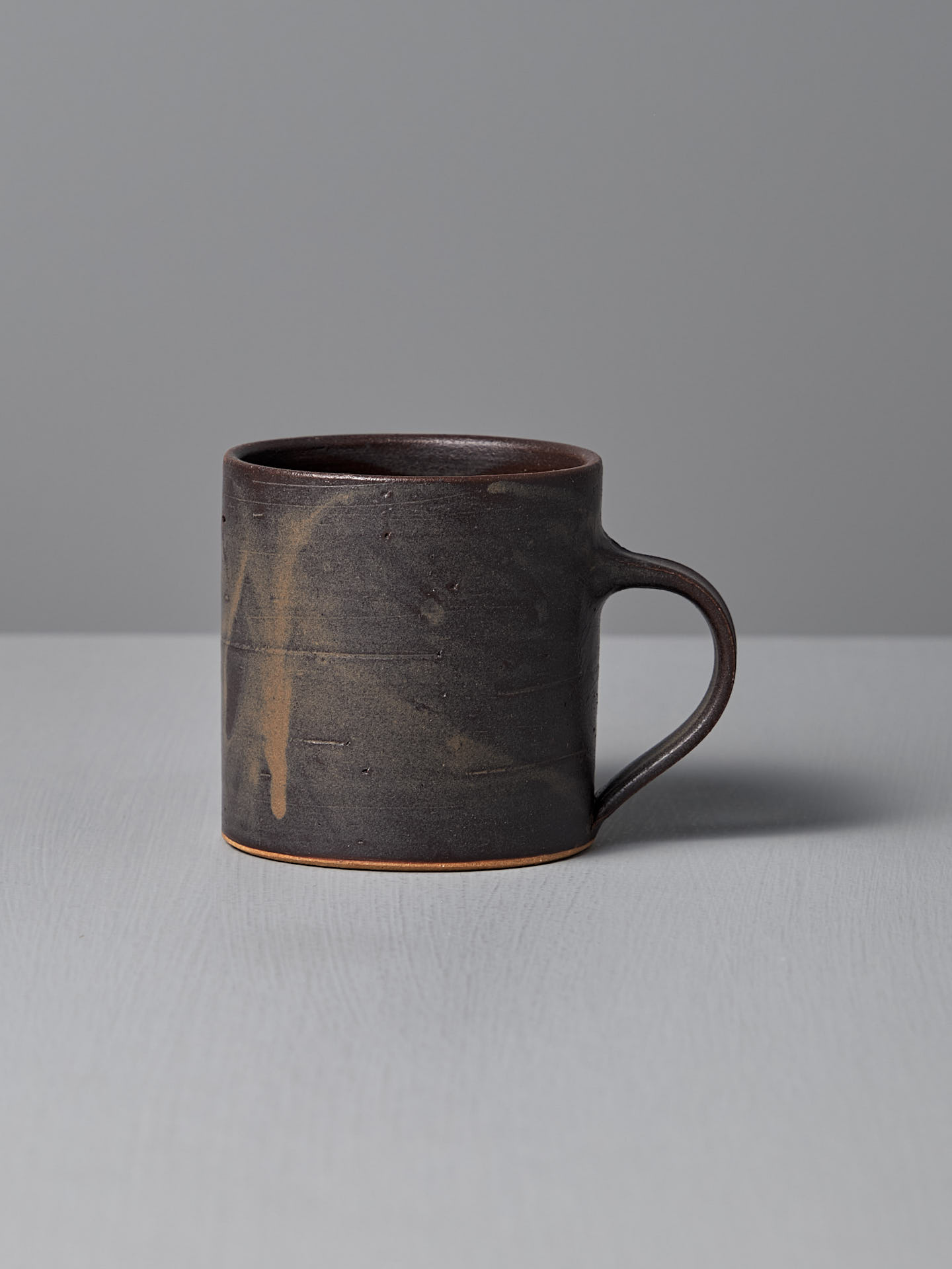 The Coffee Cup – Dapple Brown by Zoë Isaacs, a handmade mug with a subtle matte brown glaze and small handle, sits on a light gray surface against a plain gray background.